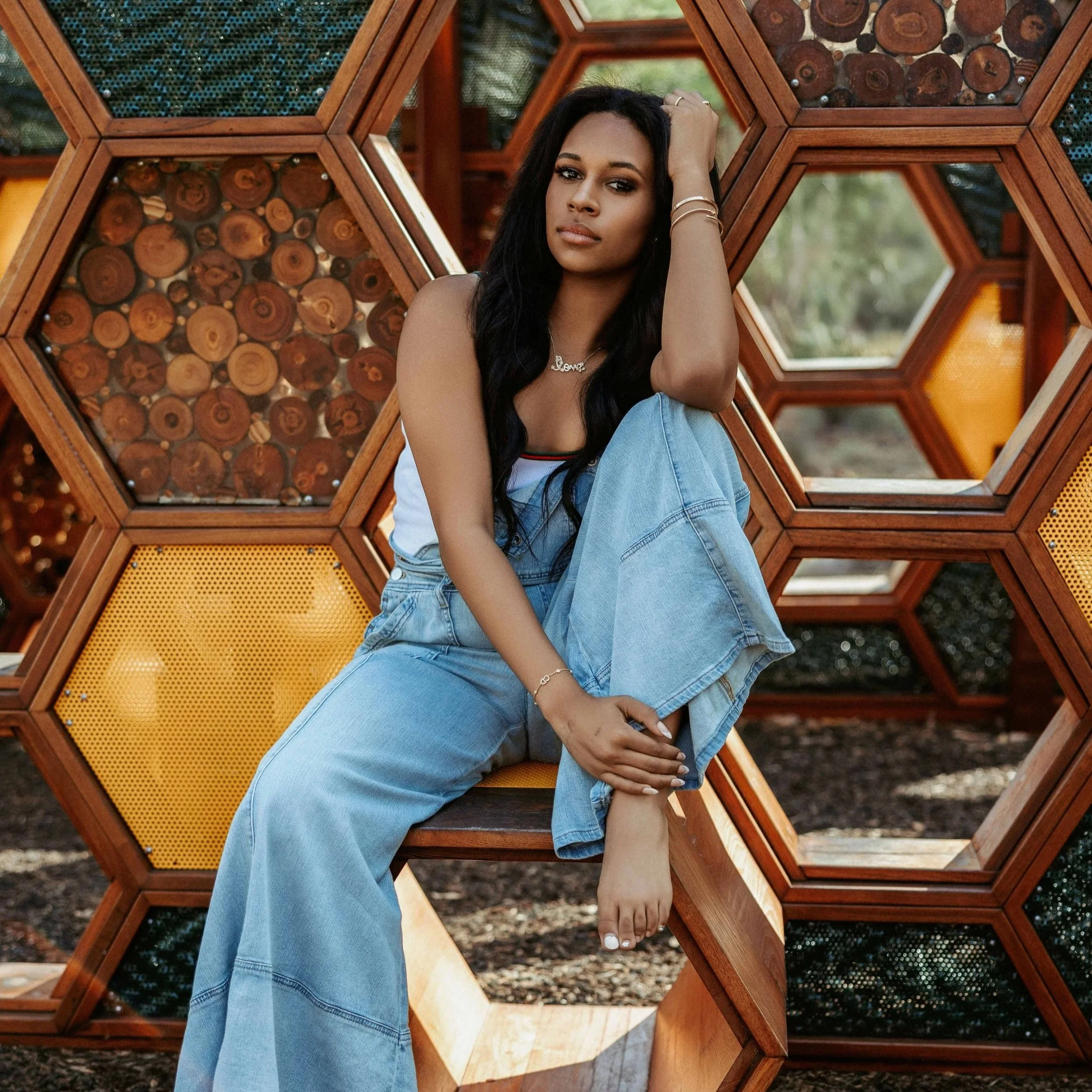 A young woman with long black hair sitting in front of a geometric wood and glass display designed with hexagonal shapes filled with wood slices and other materials, outdoors.