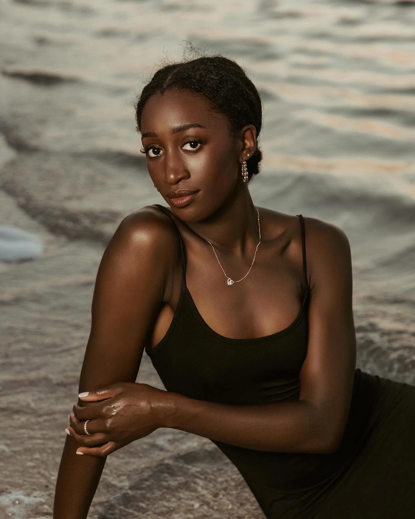 A young woman with dark skin and braided hair, wearing a black tank top, silver jewelry, and sitting on a sandy beach near the water, looking at the camera with a confident expression.