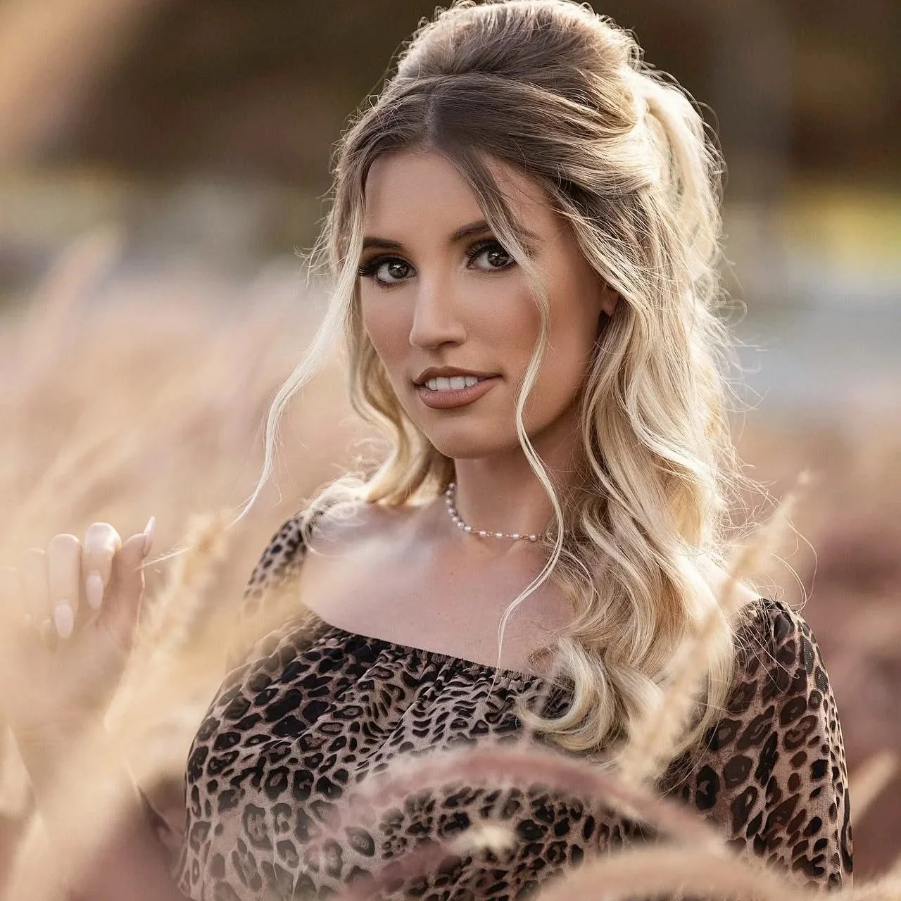 A woman with long, wavy blonde hair in a loose updo, wearing a leopard print top and a pearl necklace, standing in a field of tall grass with a blurred background.