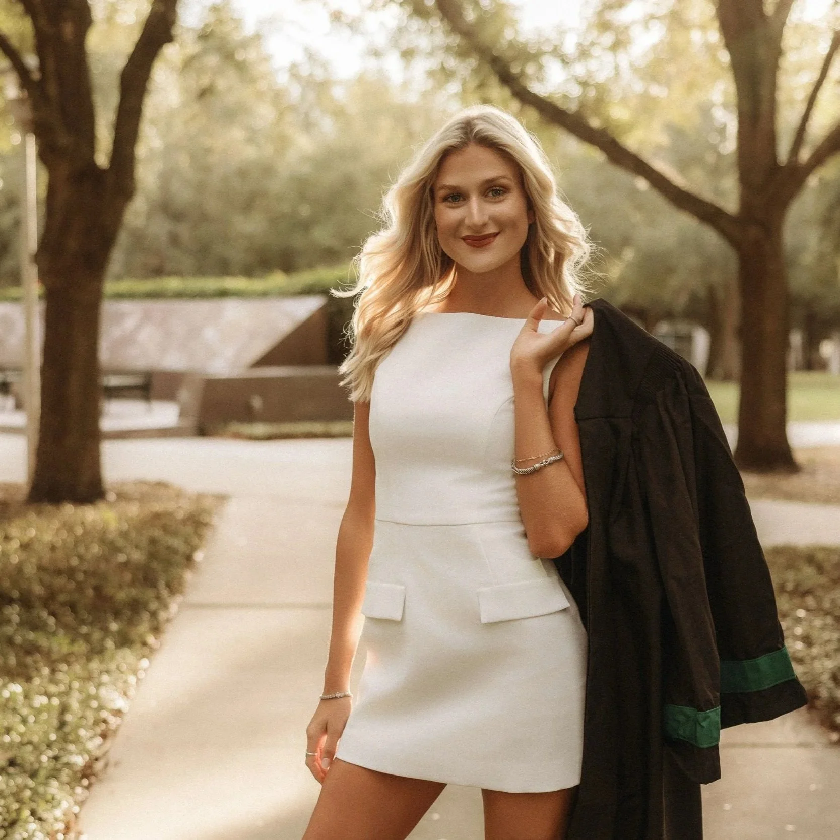 A woman with blonde hair standing outdoors in a park, wearing a white dress and holding a black jacket over her shoulder.