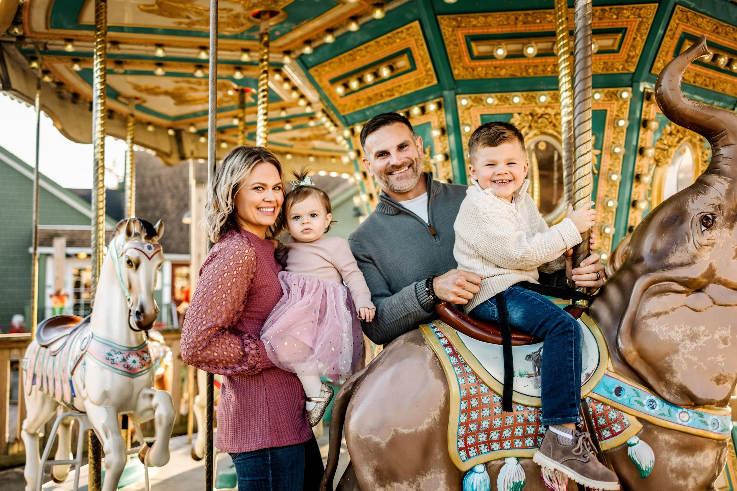 A family of four riding a carousel horse at an amusement park. The mother and father hold their young children on the carousel with a decorated horse in the foreground and a green building in the background.