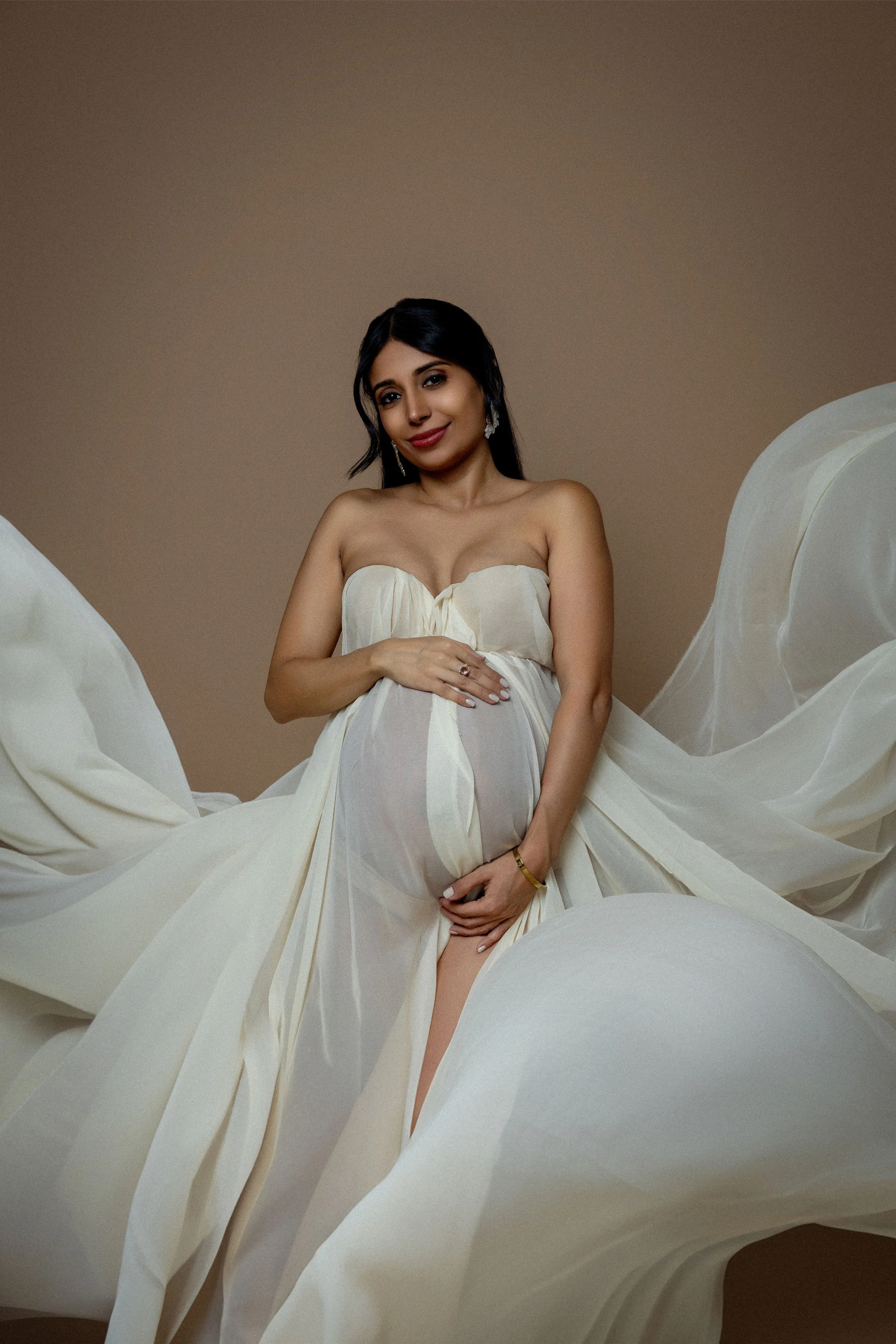 Woman wearing a strapless cream-colored gown with flowing fabric, standing against a plain background.