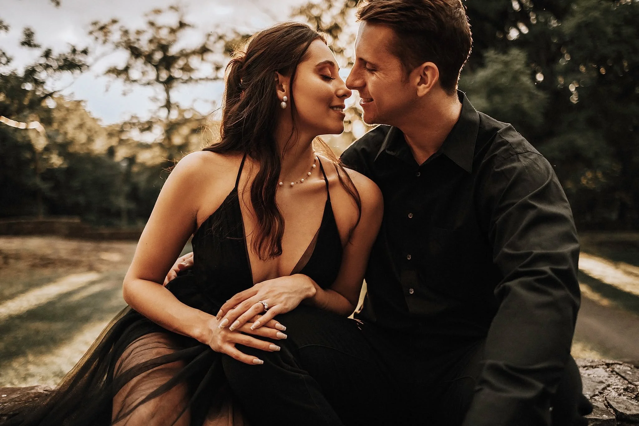A couple sitting close together outdoors, about to kiss, on a log by a lake or pond with trees in the background during sunset.