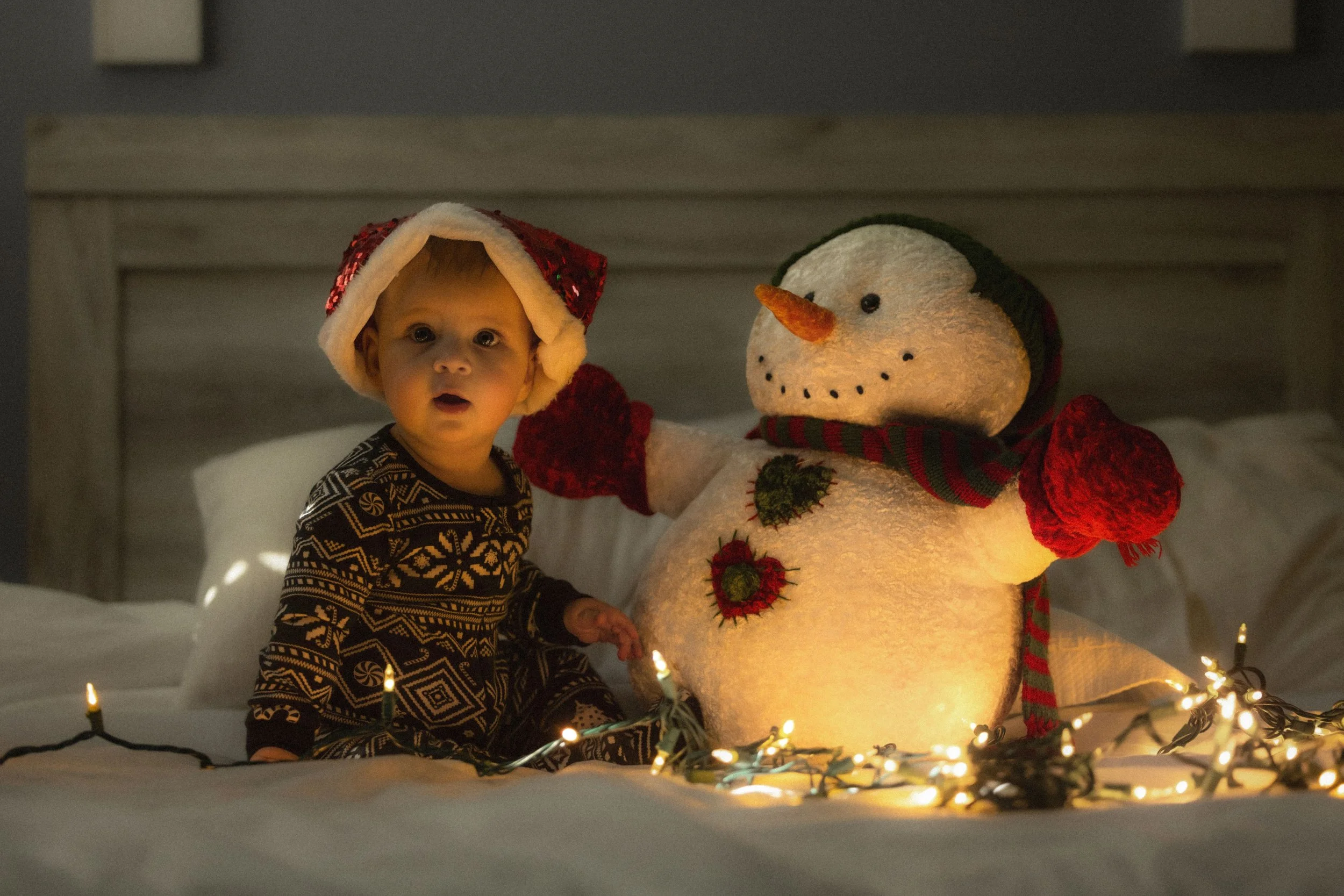 A young child wearing a Christmas-themed hat and holiday pajamas, sitting next to a lighted snowman decoration with a carrot nose, scarf, and embroidered Christmas ornaments, surrounded by holiday string lights.