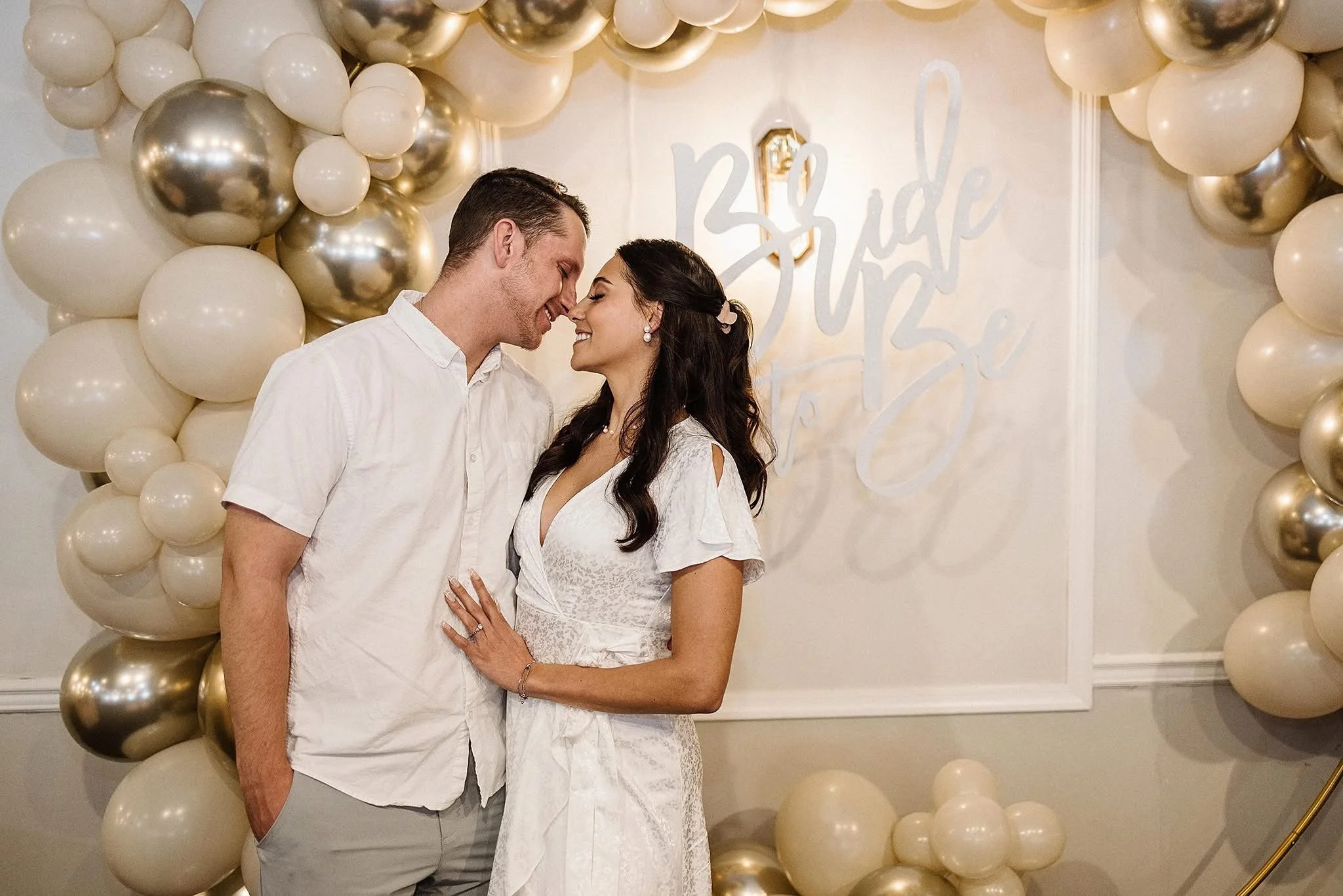 A couple standing close together, about to kiss, in front of a backdrop decorated with white and gold balloons and a sign that reads 'Bride' and 'Love', likely at a wedding celebration.