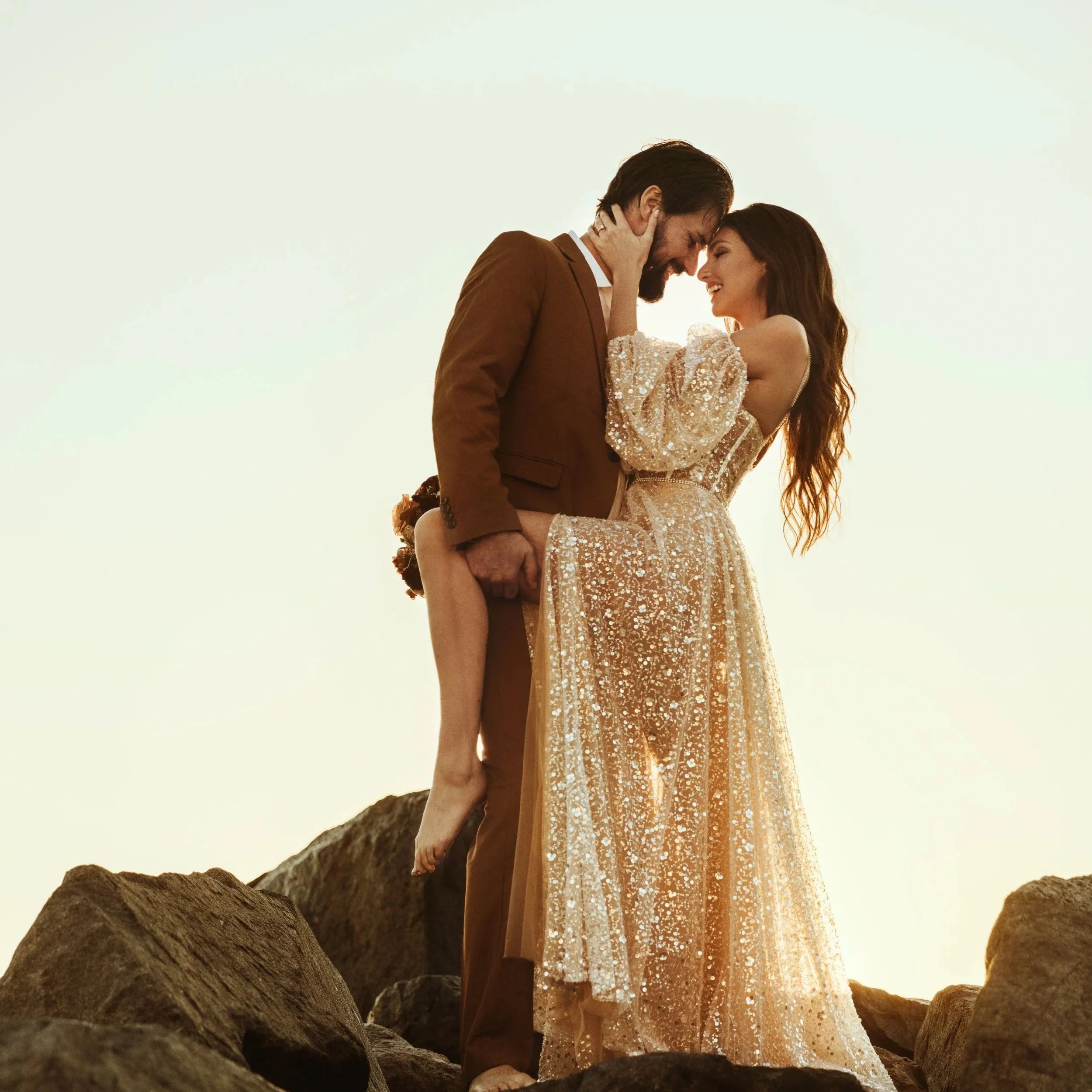A couple in vintage formal attire standing close on a rocky terrain, touching foreheads, with the sun setting or rising behind them, creating a romantic and warm atmosphere.