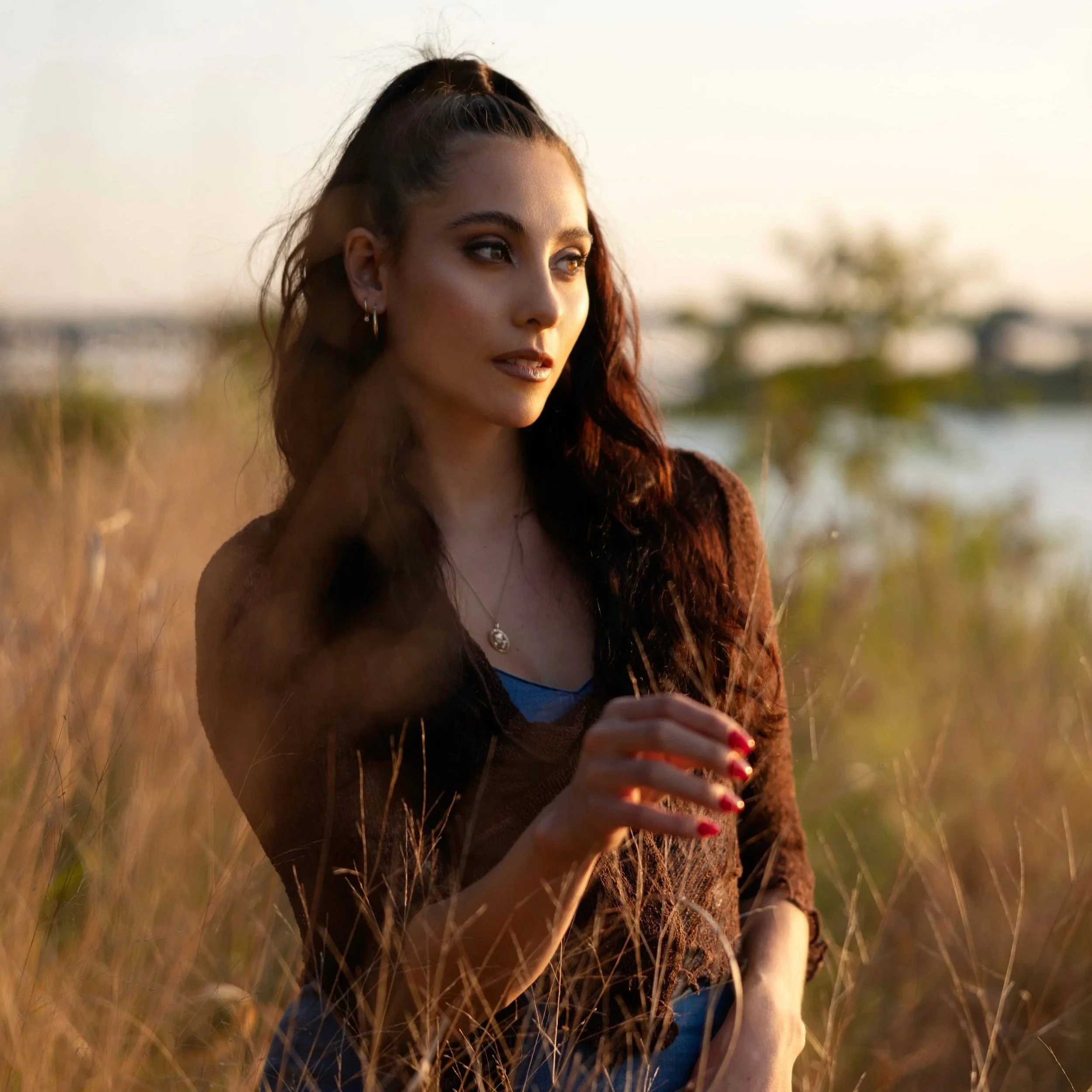 Close-up of a woman with long dark hair, some lighter streaks, wearing earrings and a necklace, standing outdoors in a field of tall grass during sunset.