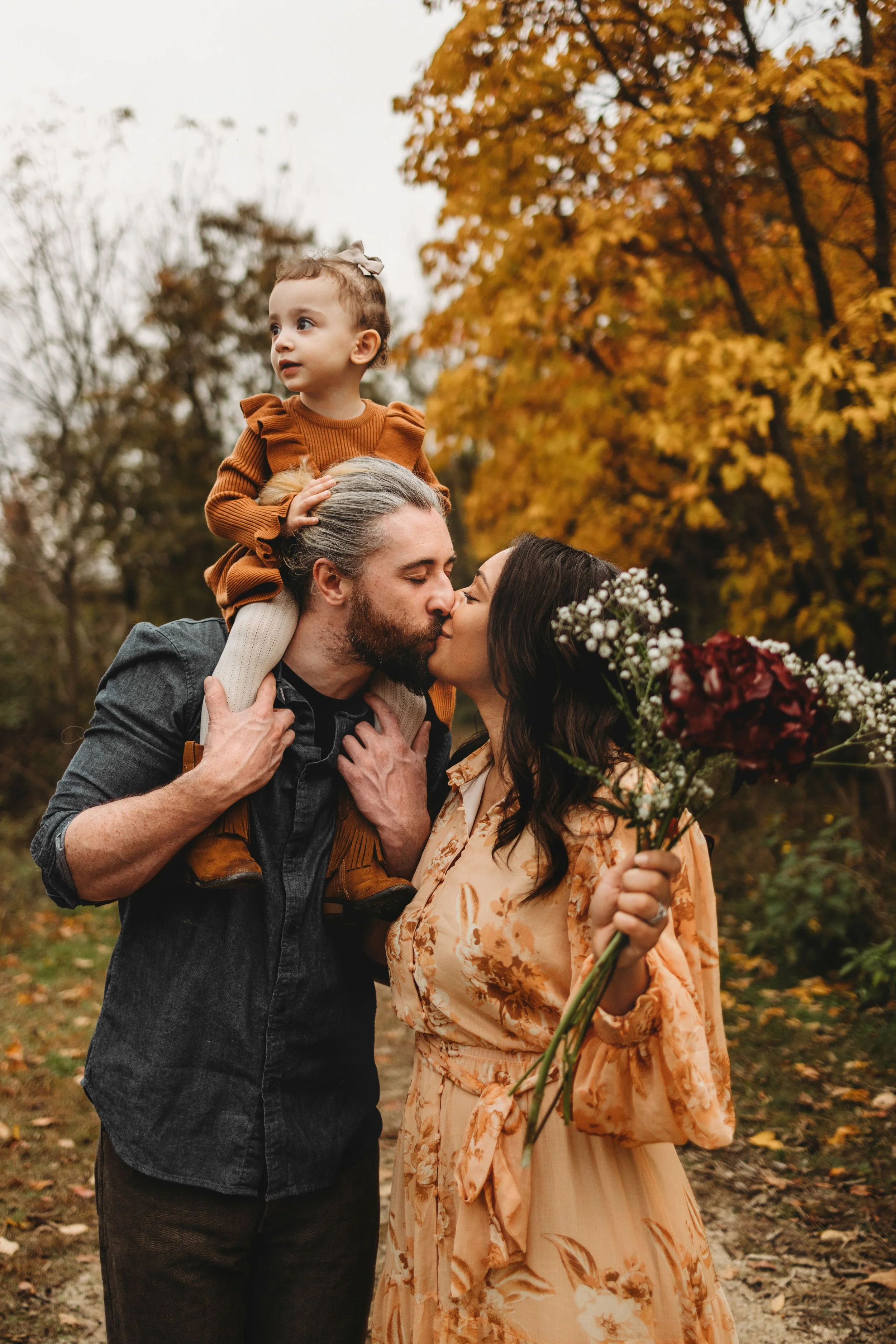 A man, woman, and young girl sharing a kiss outdoors during fall. The girl is sitting on the man's shoulders, holding his head, and the woman is holding a bouquet of flowers.