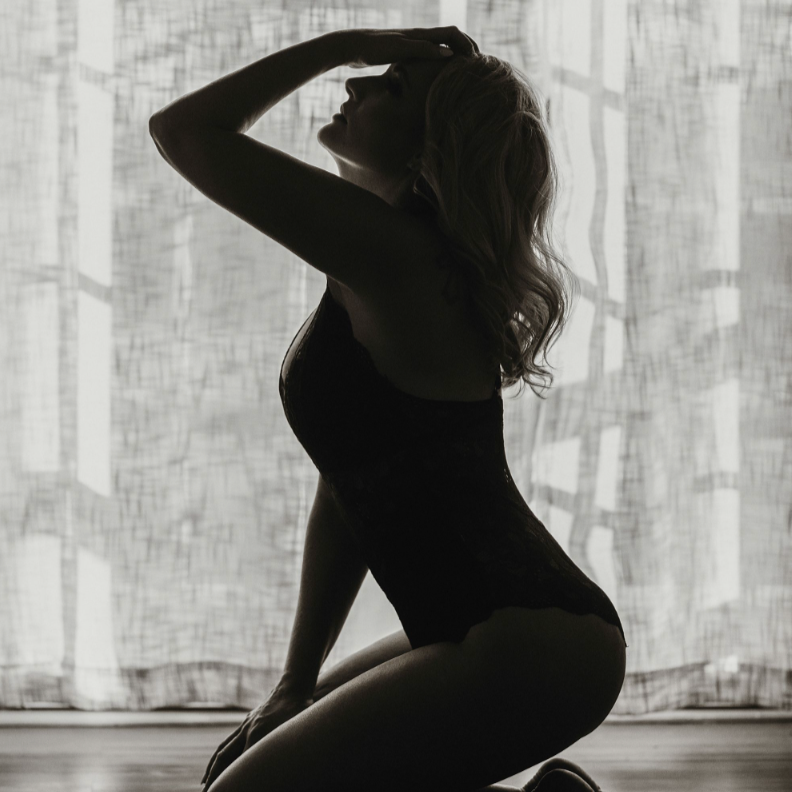 Silhouette of a woman in a black outfit kneeling with one arm raised, backlit by a patterned curtain.