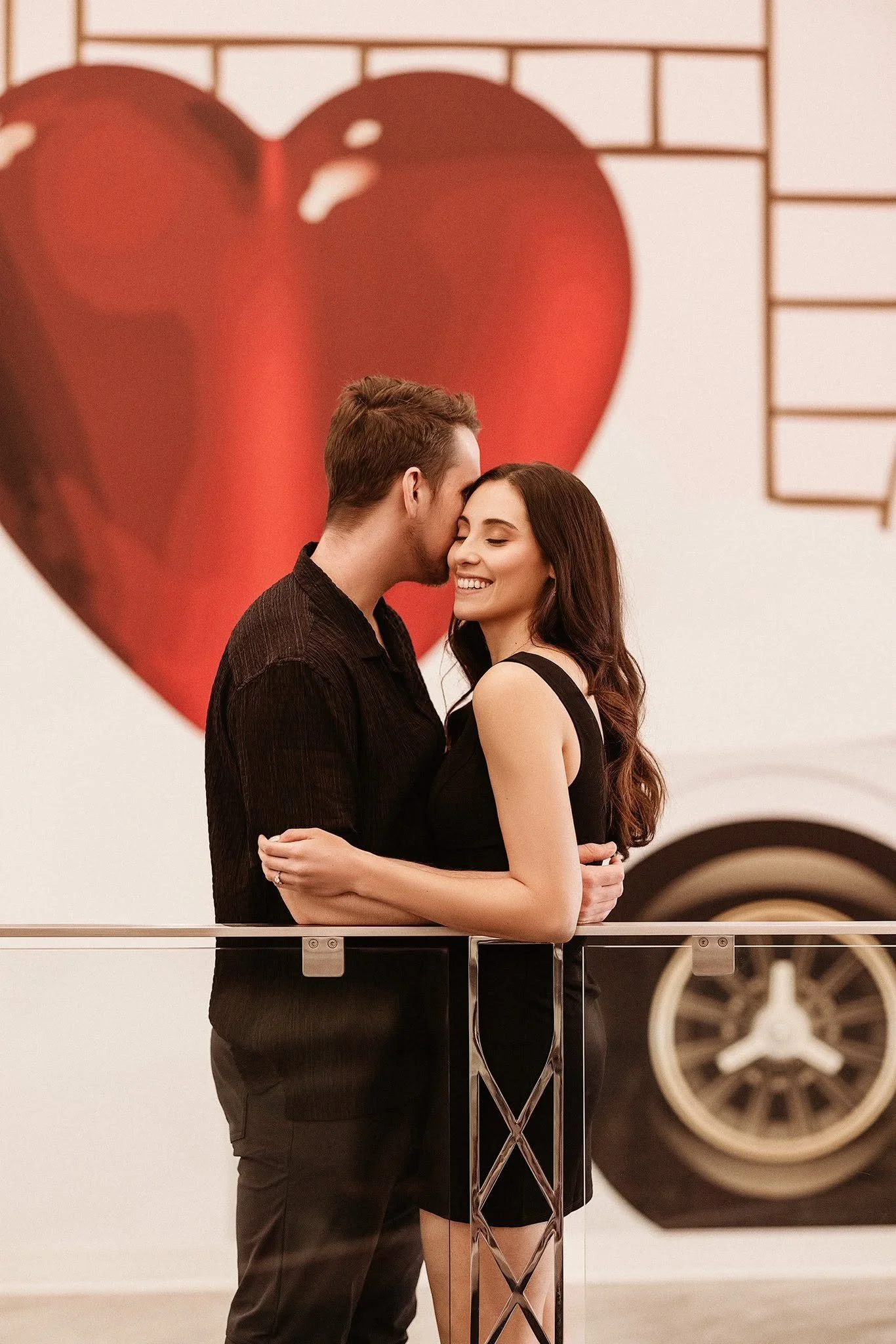 A romantic couple stands close together, smiling and embracing each other, with a large red heart artwork in the background.