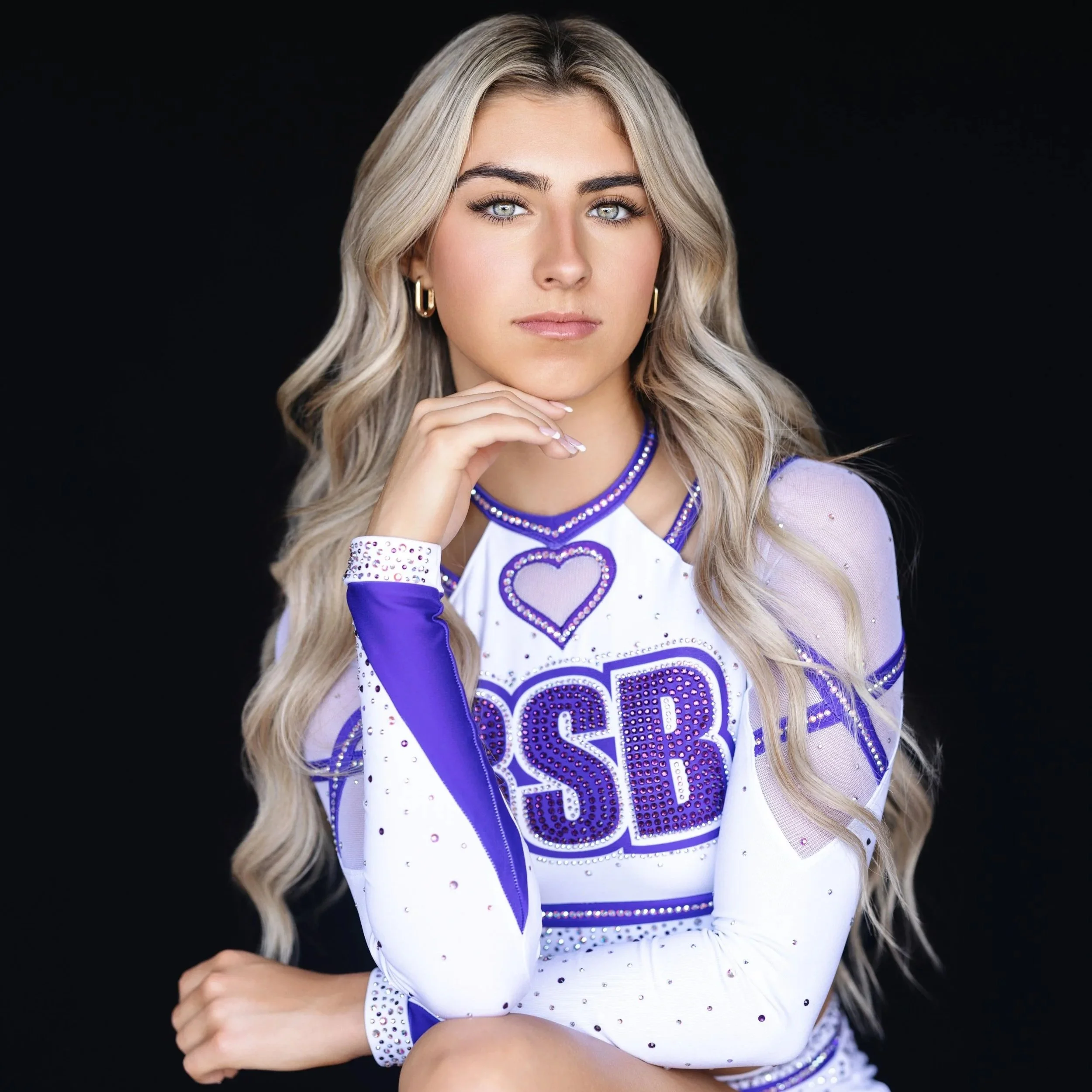 Young woman with long blonde hair and striking blue eyes posing against a black background, wearing a cheerleader outfit with purple and white accents and rhinestone embellishments, resting her chin on her hand.