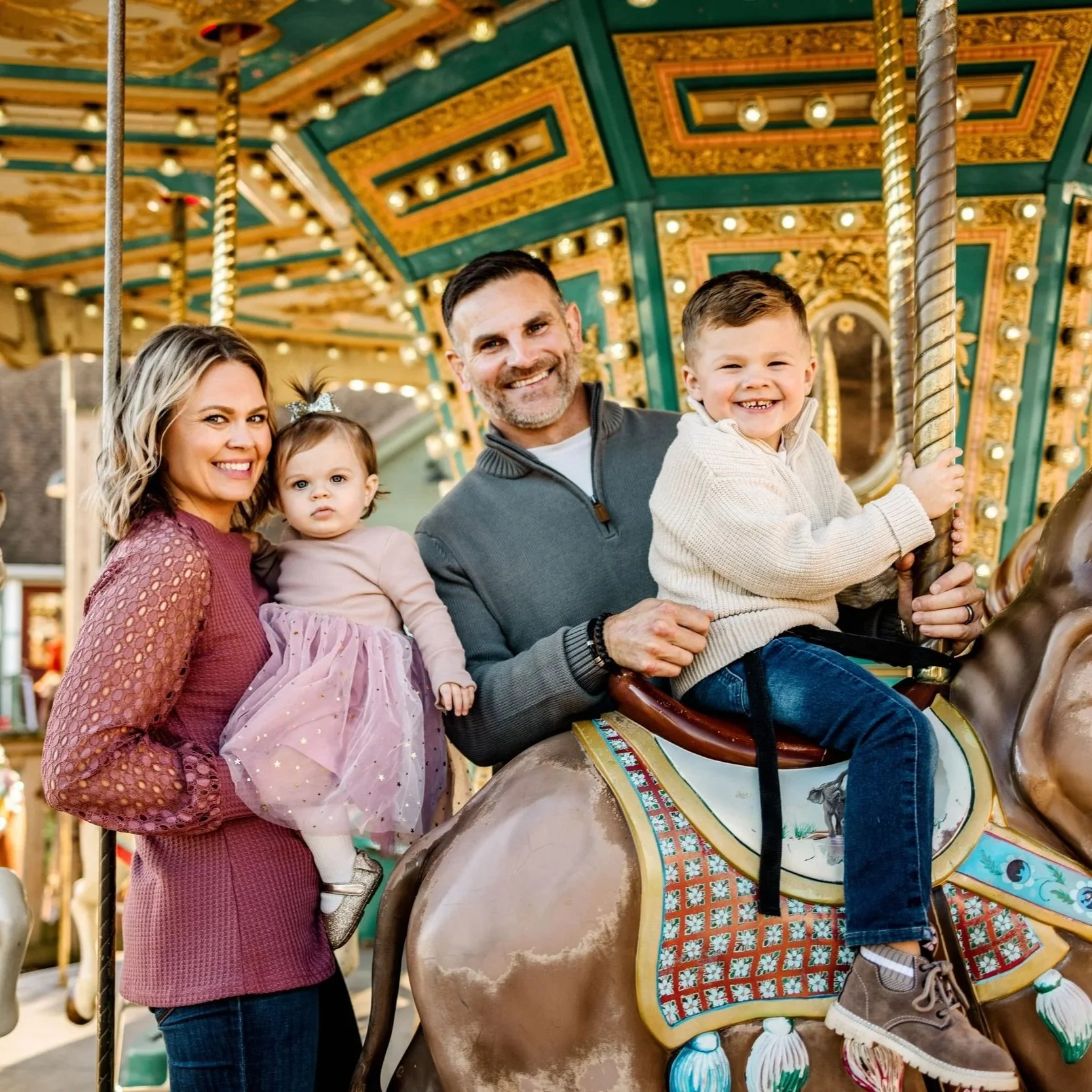 A smiling family of four riding a carousel horse at an amusement park. The woman and two children sit on the horse, while the man holds the child in front of him. The carousel is decorated with colorful patterns and lights.