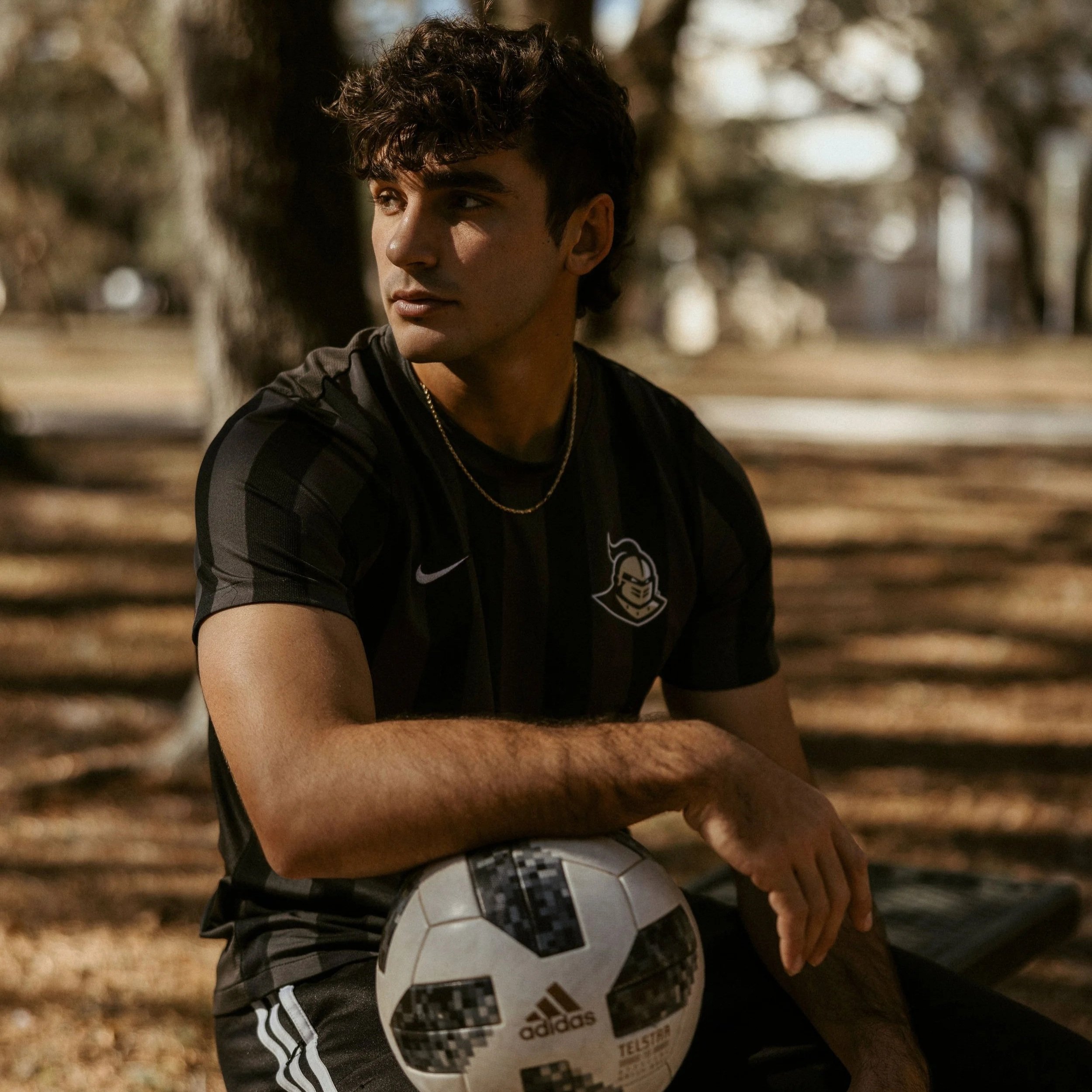 A young man with dark curly hair and light brown skin wearing a black Adidas sports shirt holding a soccer ball with a black, white, and grey pattern, outdoors with blurred trees in the background.
