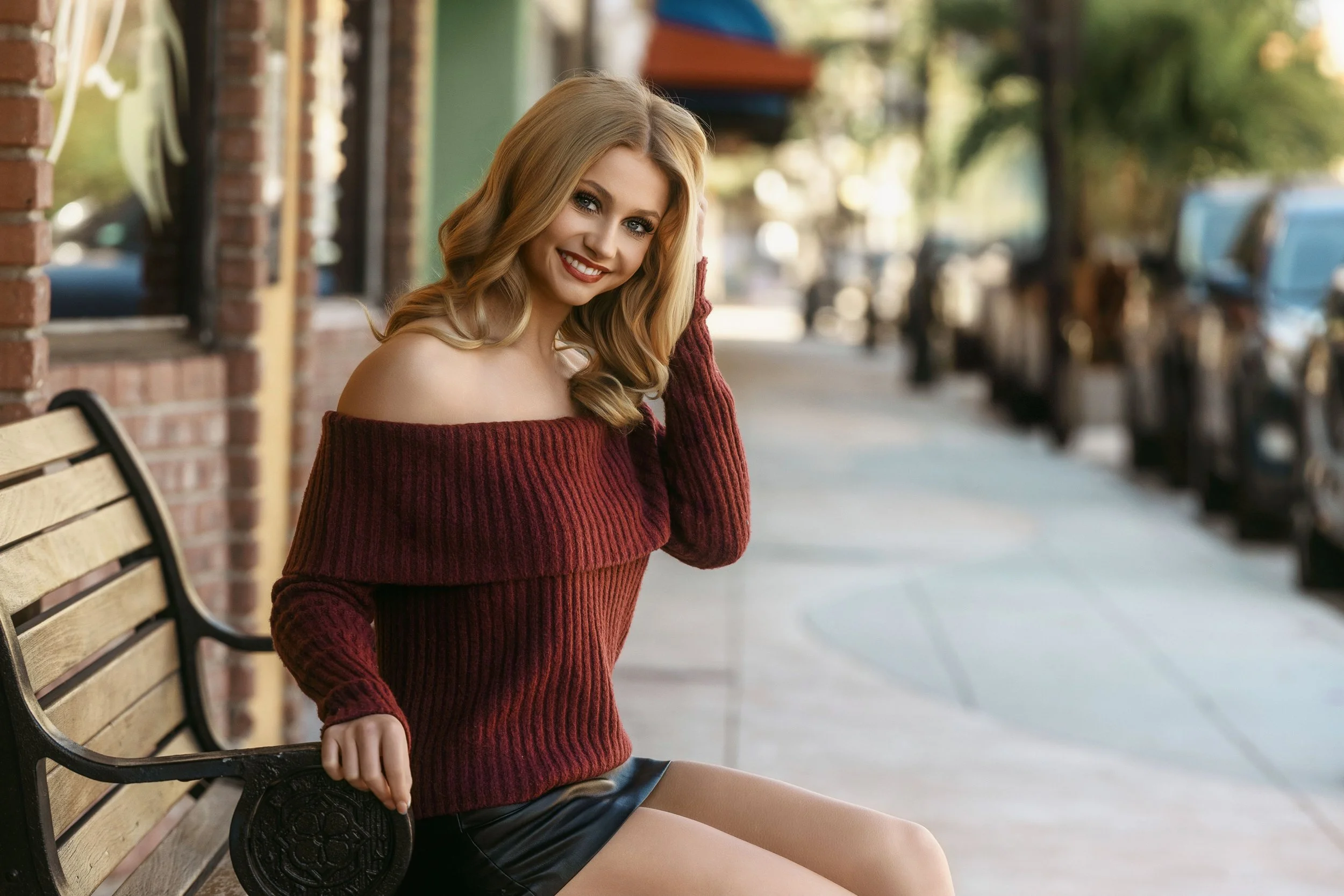 A smiling young woman sitting on a bench outdoors, wearing a red off-the-shoulder sweater and black shorts, with her hand resting on her head, in a park or street setting with trees and parked cars in the background.