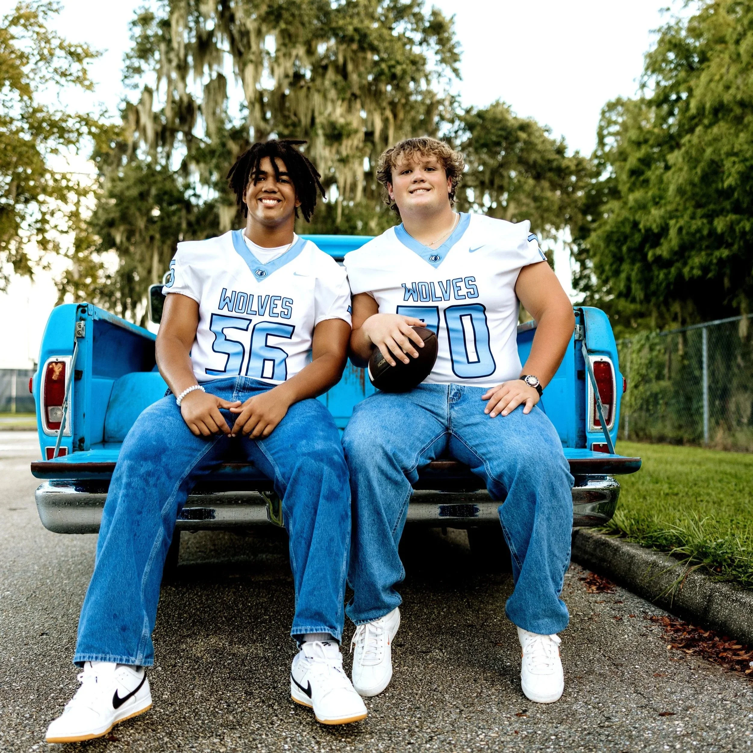Two young men wearing white football uniforms with blue accents and the word "Wolves" on the front, sitting on a blue bench with their legs crossed, in an outdoor park setting with green trees in the background.