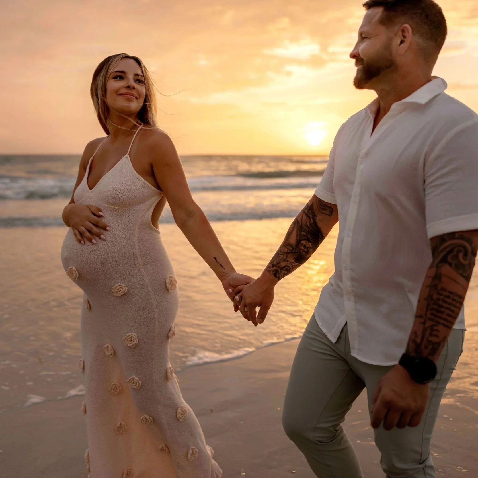 A pregnant woman and a man holding hands on the beach during sunset.