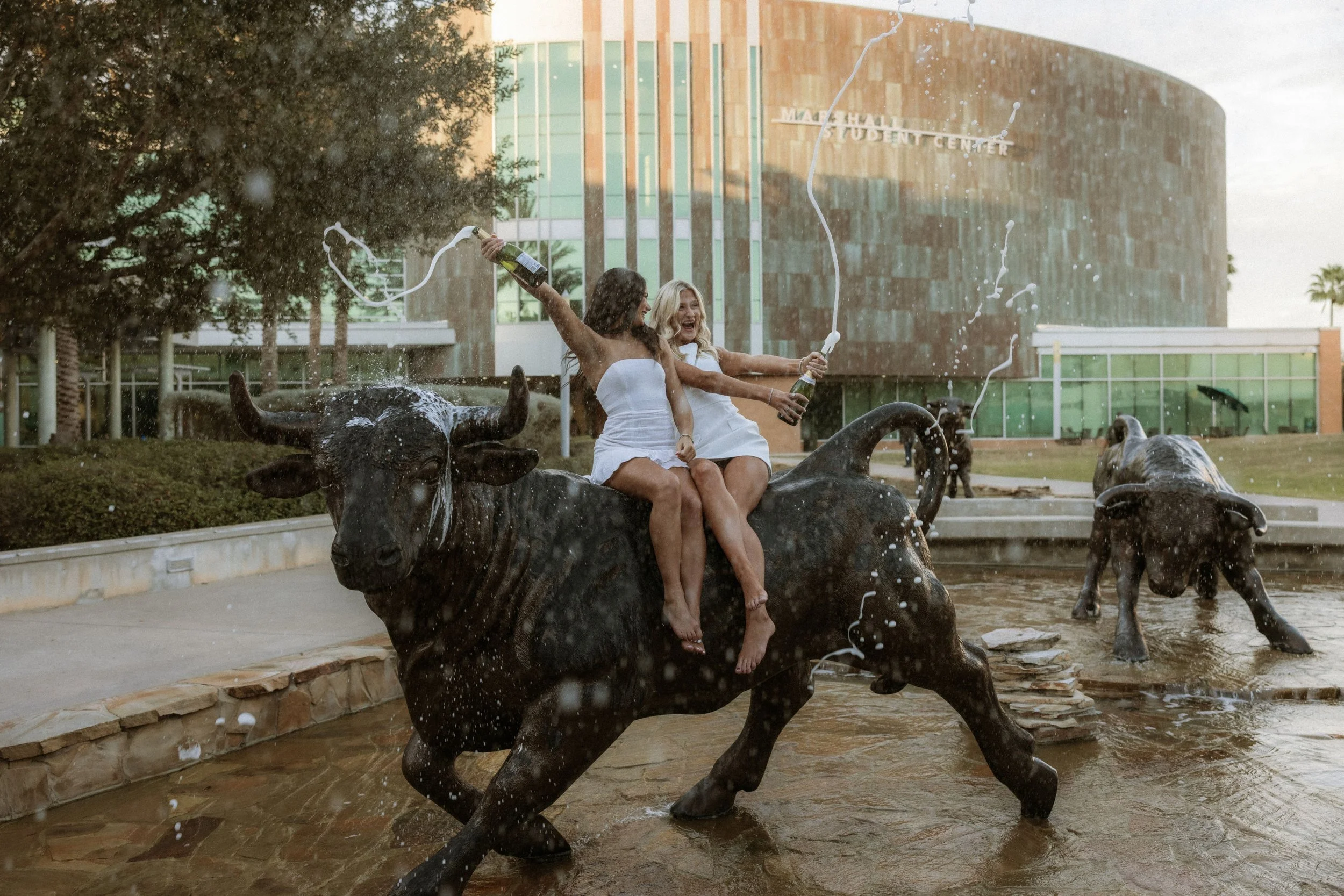 Two women in white dresses sitting on a bronze cow statue in a fountain, splashing champagne, with a modern building and trees in the background.