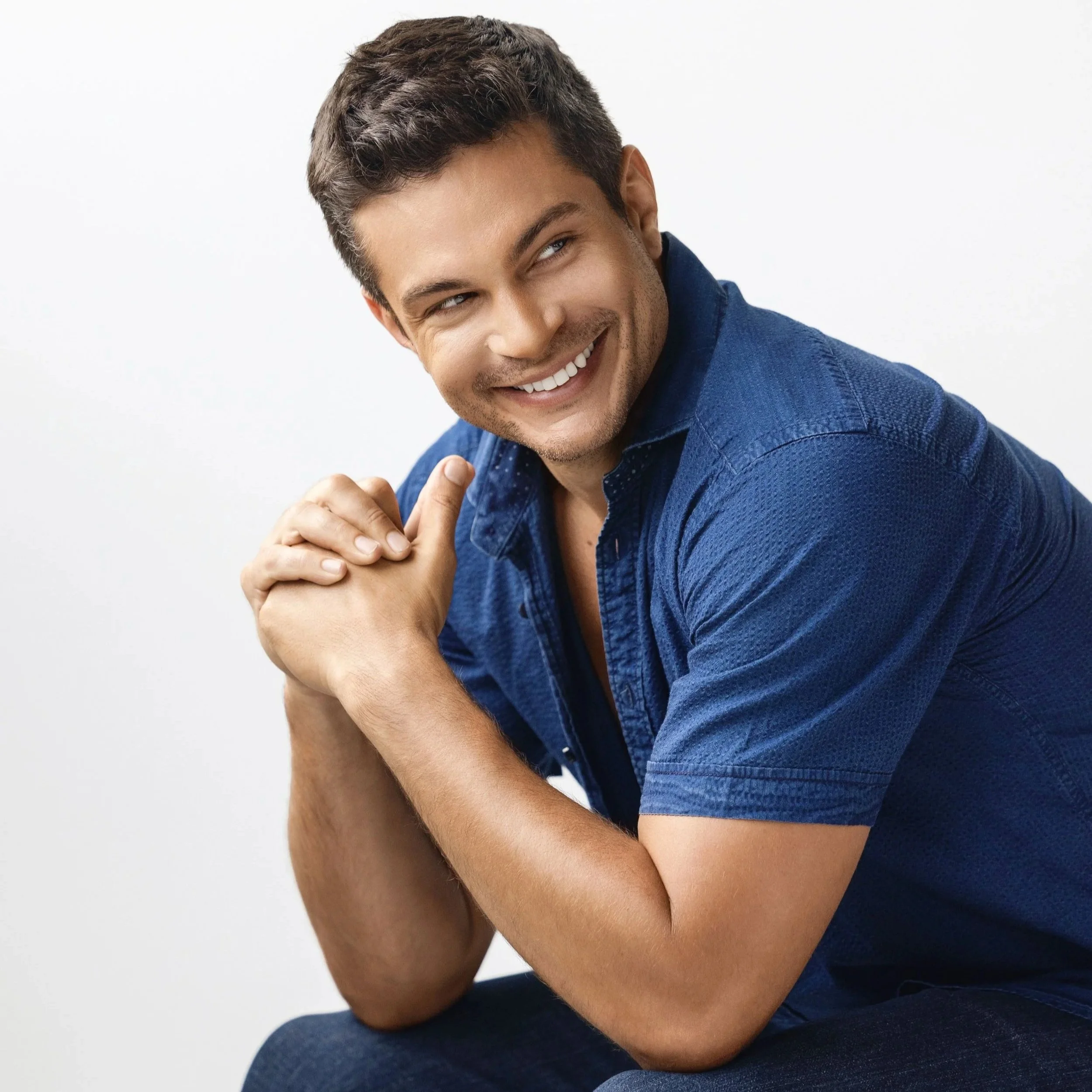 A young man with dark hair styled upward, smiling and looking to his right, wearing a blue shirt, with his arms crossed and hands on his shoulders, against a plain white background.