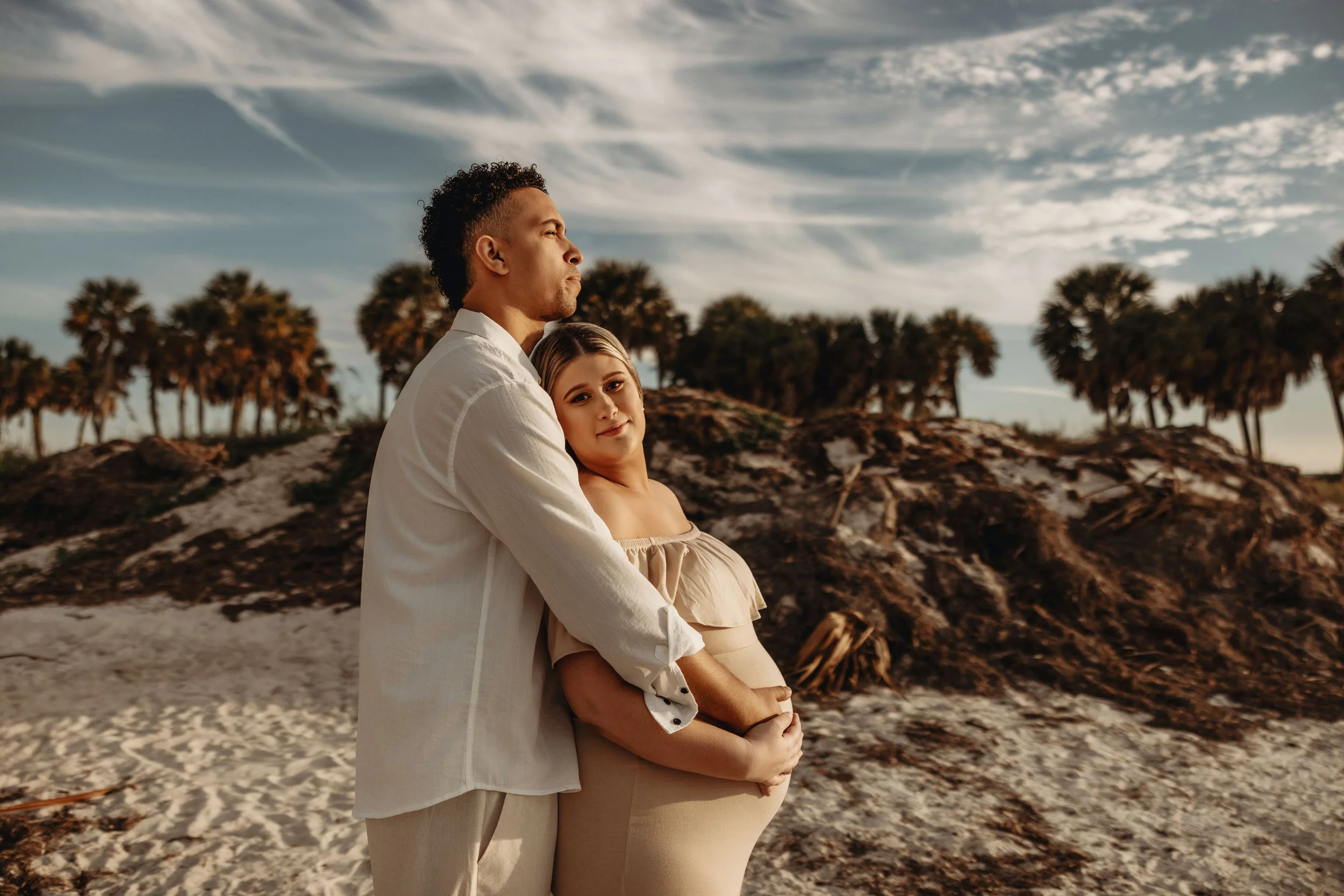 A couple embracing on a beach at sunset or late afternoon, with palm trees in the background and an expansive sky with wispy clouds.
