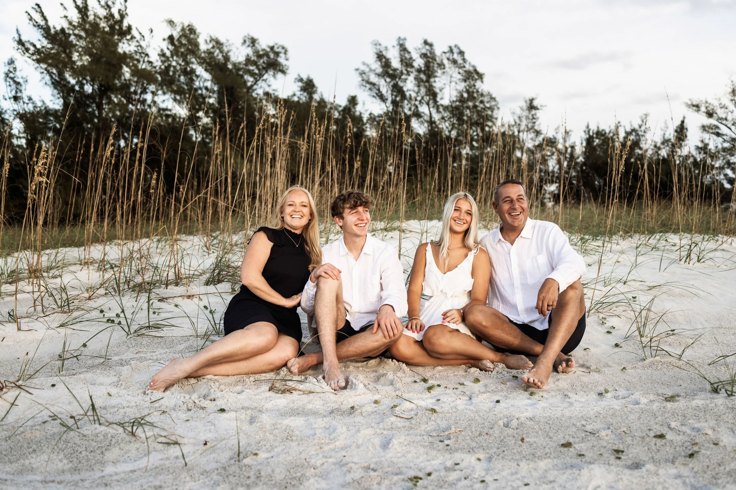 Family sitting on the sand at the beach, smiling with dune grass and trees in the background.