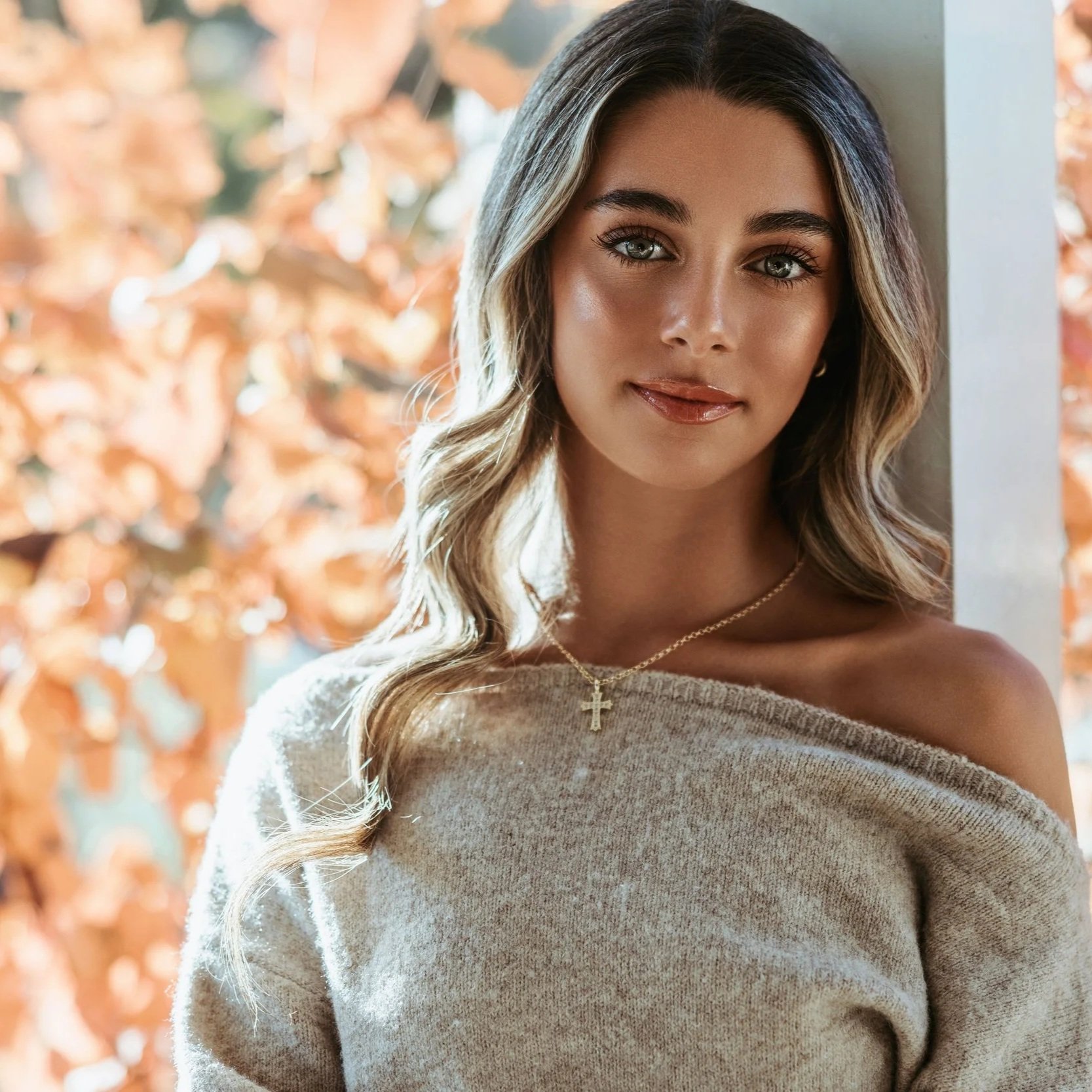 Close-up photo of a woman with long wavy hair, wearing a light-colored sweater, a gold cross necklace, and hoop earrings, standing in front of an autumn background.
