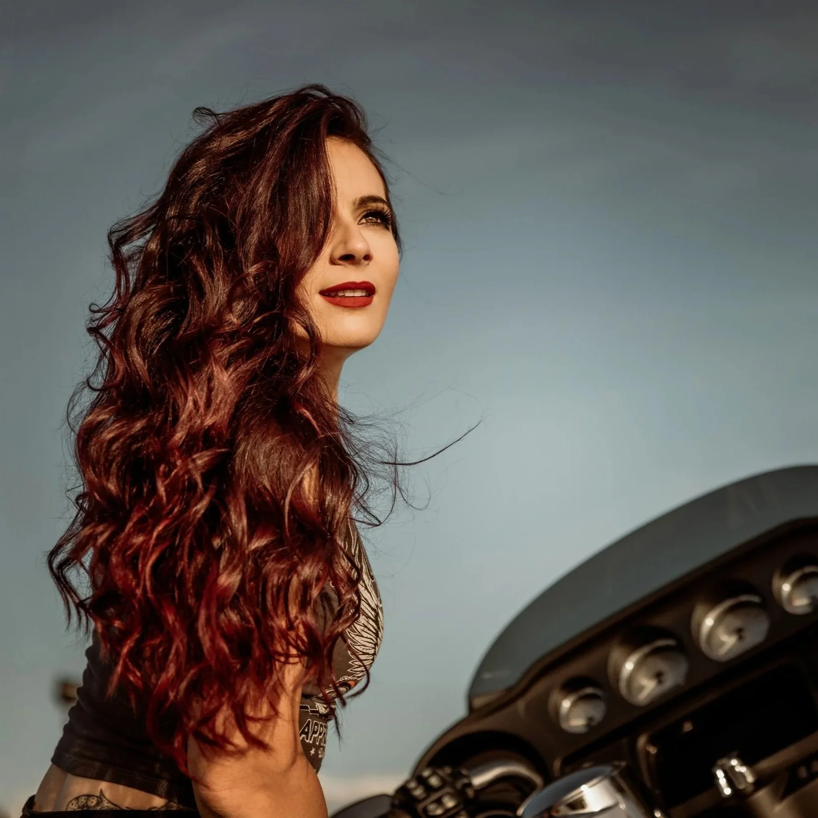 A woman with long curly red hair looking up, standing in front of a black light fixture with multiple circular bulbs. She is wearing makeup with red lipstick and a patterned top.