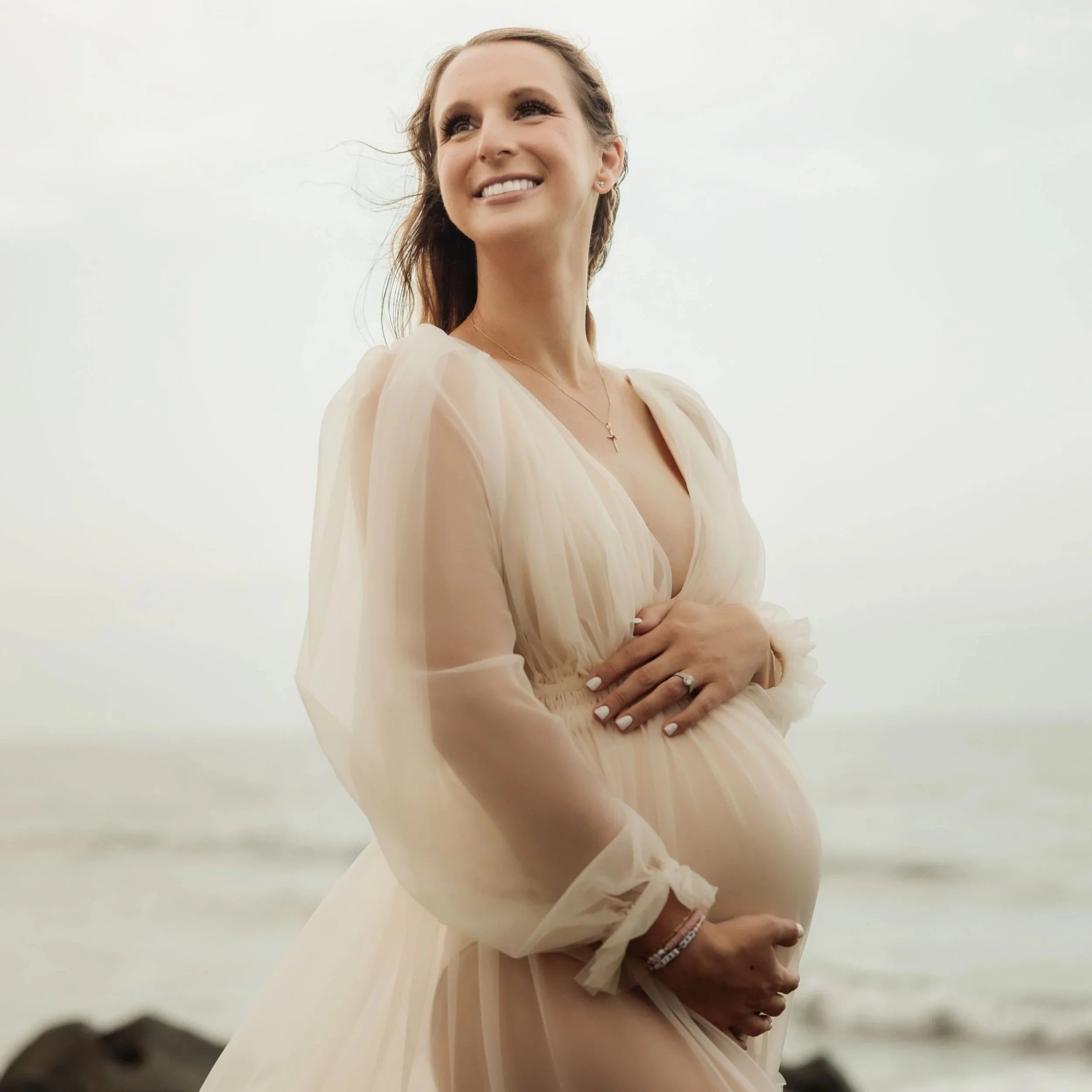A woman with a smile, wearing jewelry and a light, elegant dress, standing outdoors against a pale sky.
