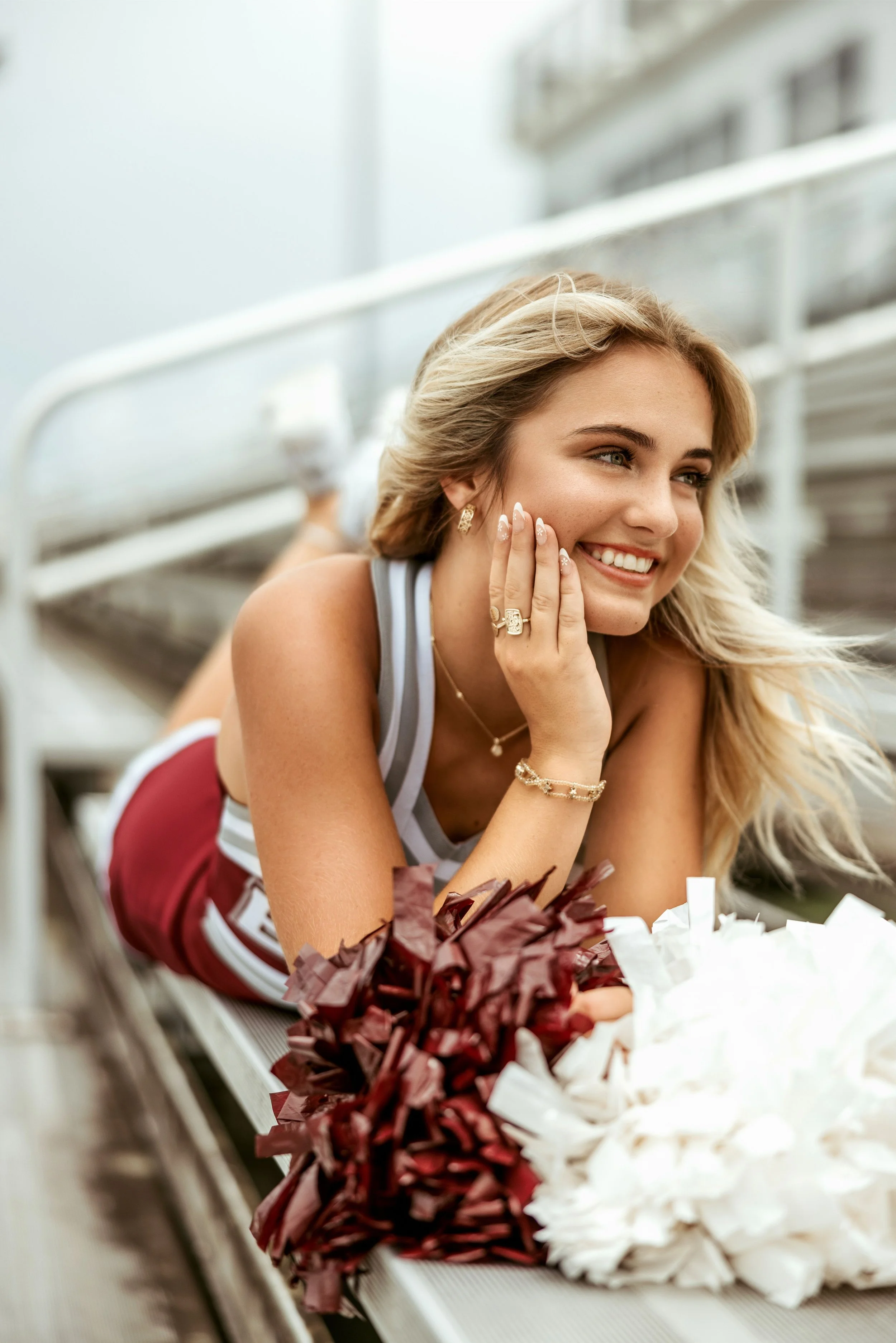 Smiling young woman lying on her stomach on a bench at a stadium, with pom-poms in front of her, resting her face on her hand, wearing a cheerleading uniform and jewelry.