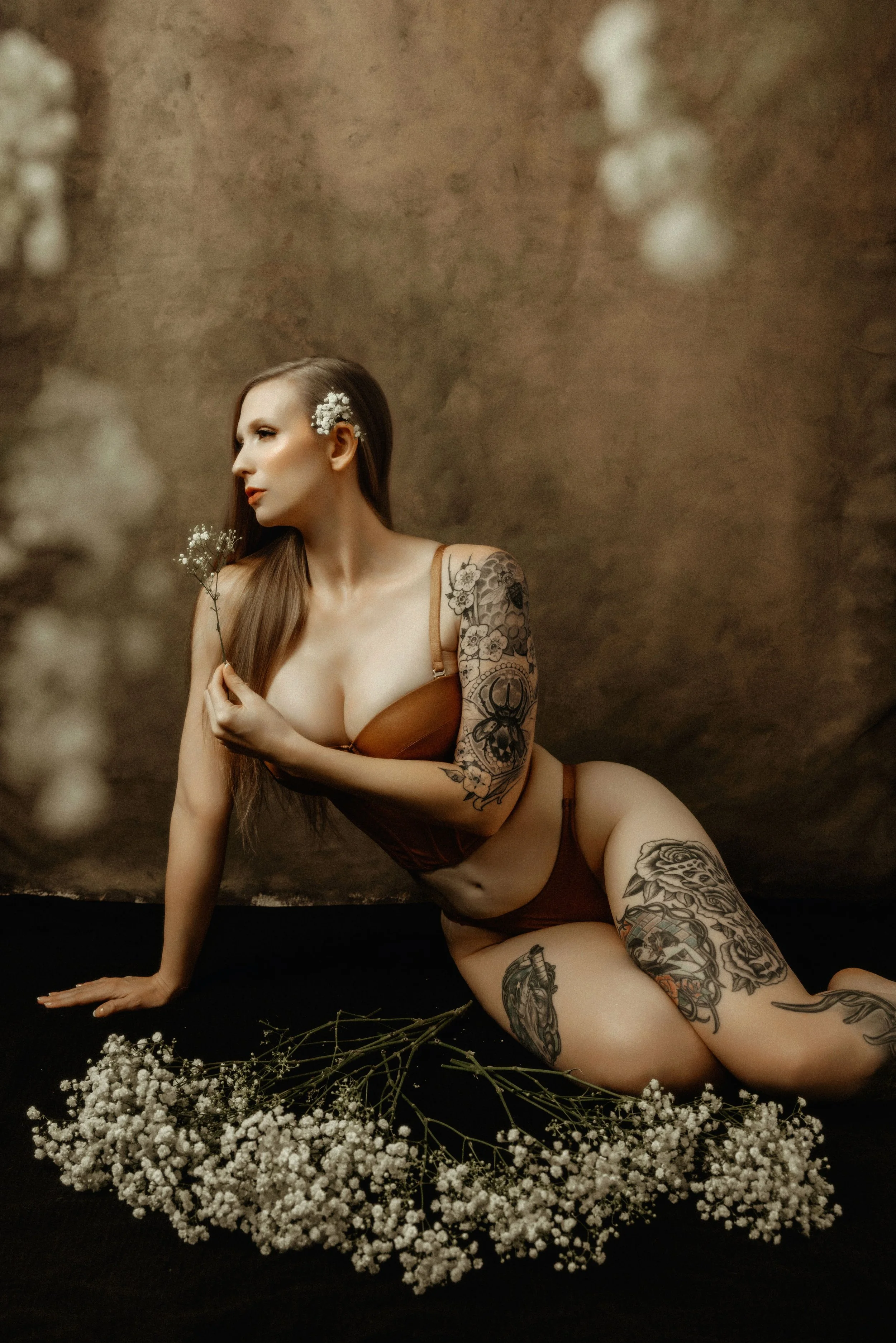 A woman with long brown hair and tattoos, dressed in brown lingerie, is sitting on a black surface surrounded by white baby's breath flowers. She has white flowers in her hair and is holding a small sprig of flowers, with a neutral expression on her face.