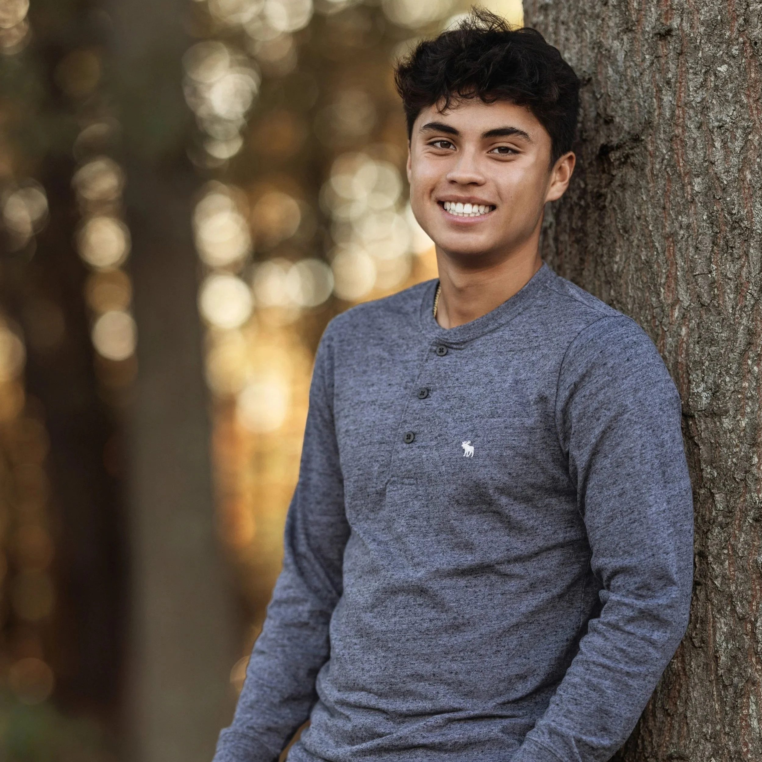 A young man with dark, curly hair, smiling showing his teeth, wearing a gray button-up shirt, leaning against a large tree trunk outdoors during sunset or golden hour.