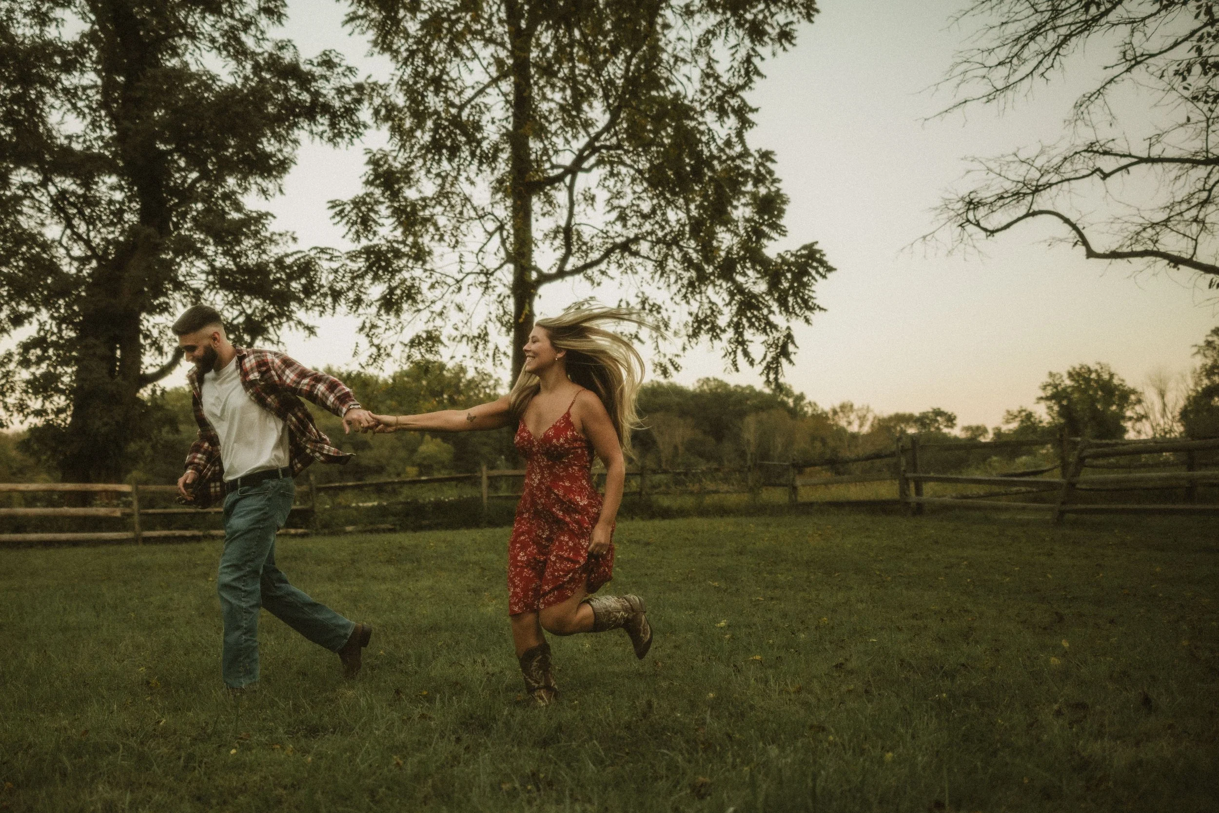 A couple holding hands and running through a grassy field with trees in the background during sunset.