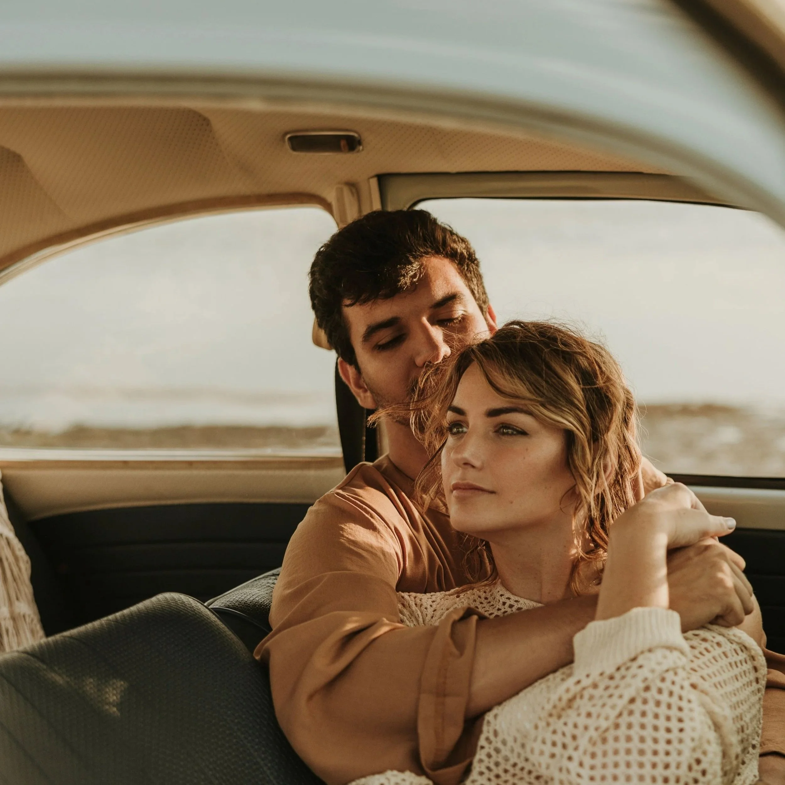 A young man and woman sit close together in a car, embracing affectionately. The woman's hair is reddish-brown and she wears a white crocheted top, while the man has dark curly hair. They are looking at each other lovingly, with a scenic landscape visible through the car window in the background.