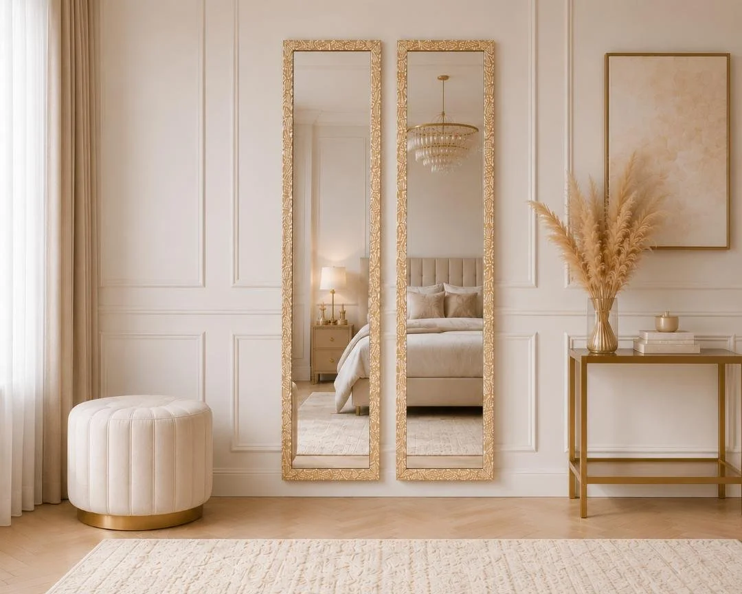 Elegant bedroom with a white and gold color scheme, featuring two tall ornate gold framed mirrors, a beige upholstered bed with pillows, a side table with a lamp, a dresser with a vase of pampas grass, and abstract artwork.