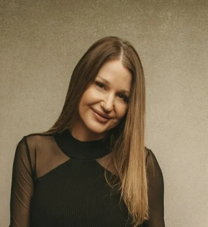 A woman with long brown hair smiling and wearing a black top with sheer sleeves, standing against a beige background.
