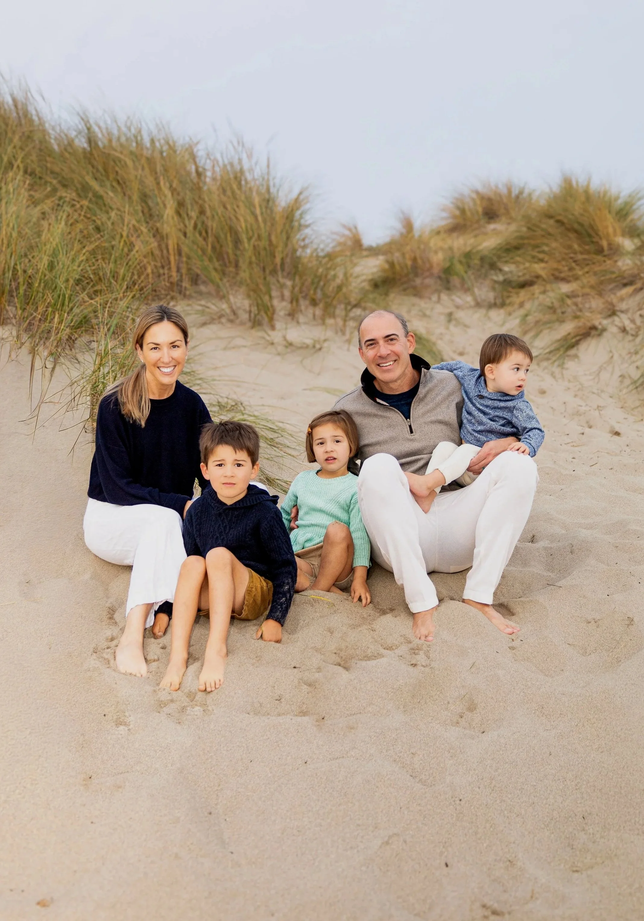 Family of five sitting in the sand on a beach with dunes and grass in the background, smiling at the camera.