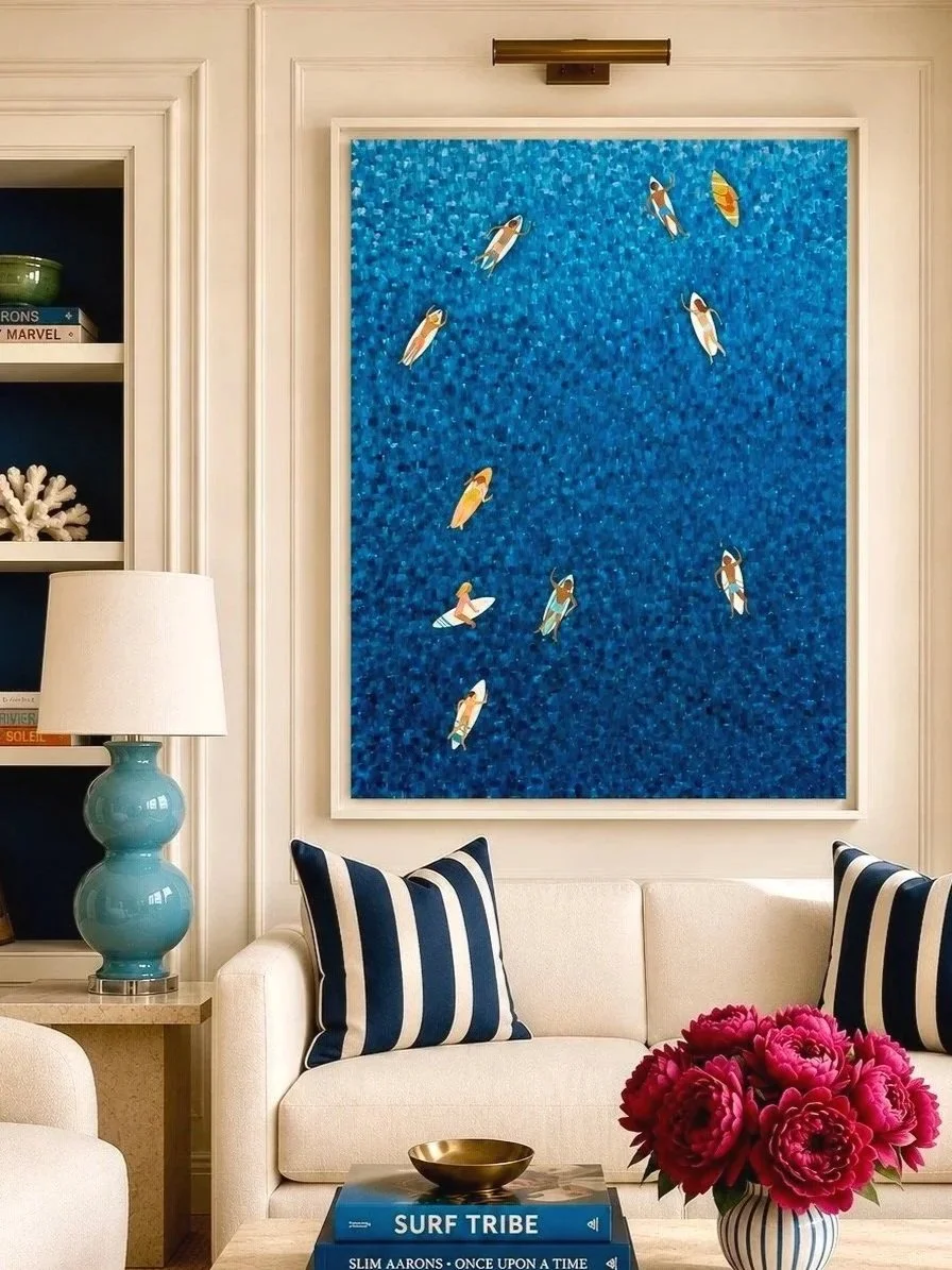 Living room with a large blue painting of tiny surfers on the wall, a white sofa with navy and white striped pillows, a blue ceramic table lamp, a side table with pink peonies, and books titled 'Surf Tribe' and 'Slim Aarons' on a coffee table.
