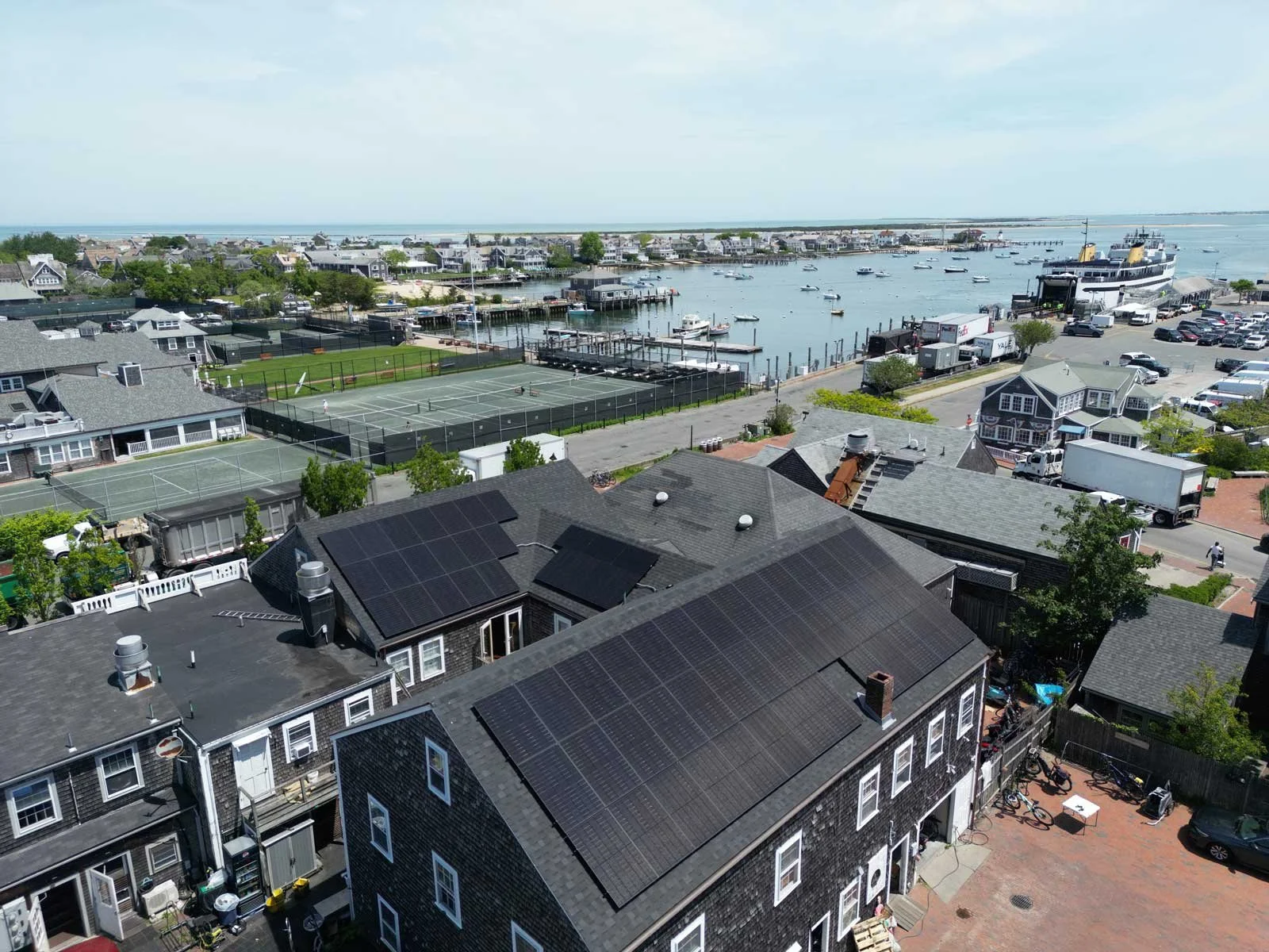 Aerial view of a coastal town showing rooftops with solar panels, tennis courts, a marina with boats, and the ocean in the background.