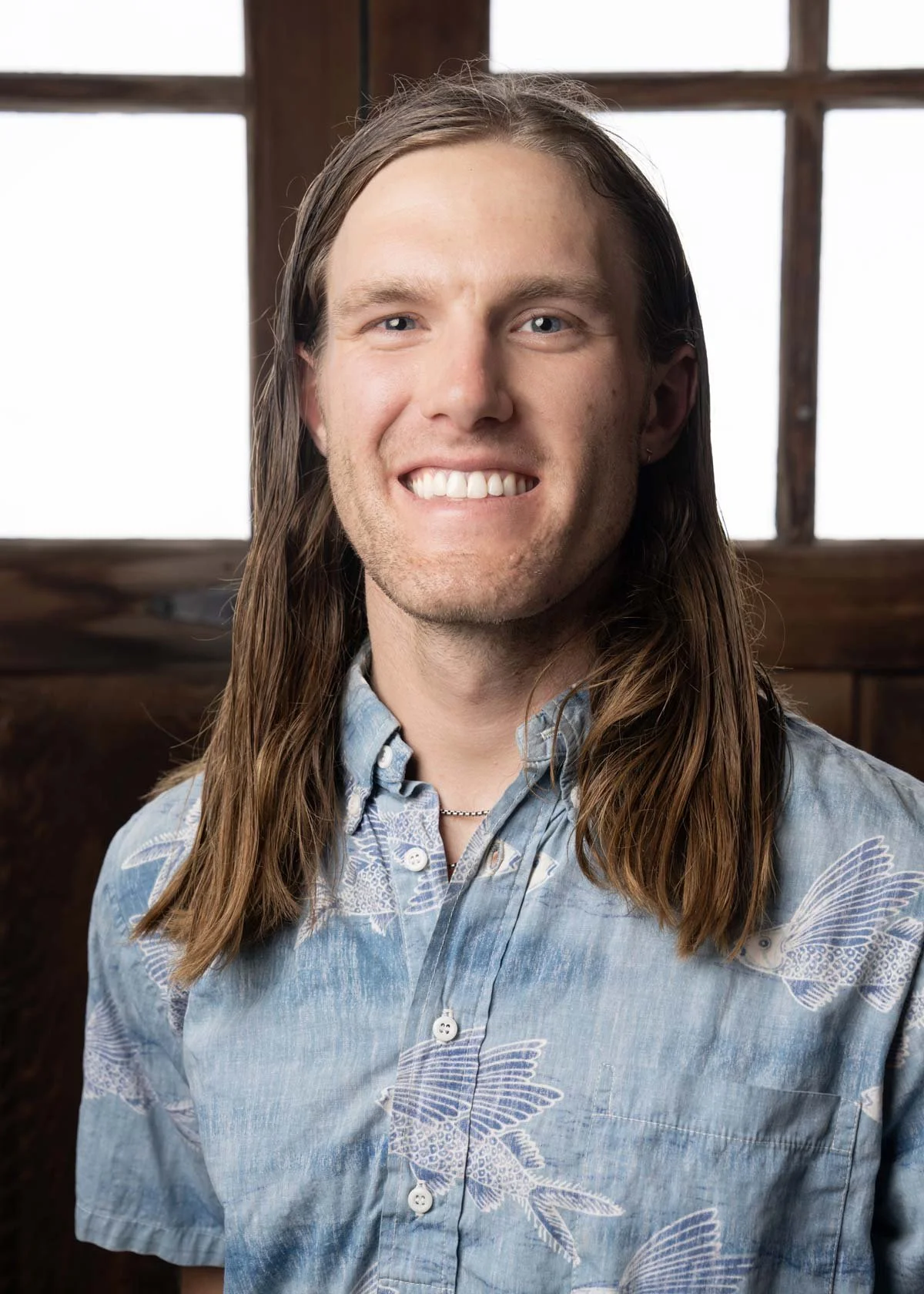 A young man with long brown hair, smiling with teeth visible, wearing a blue patterned button-up shirt, standing in front of a wooden window frame.