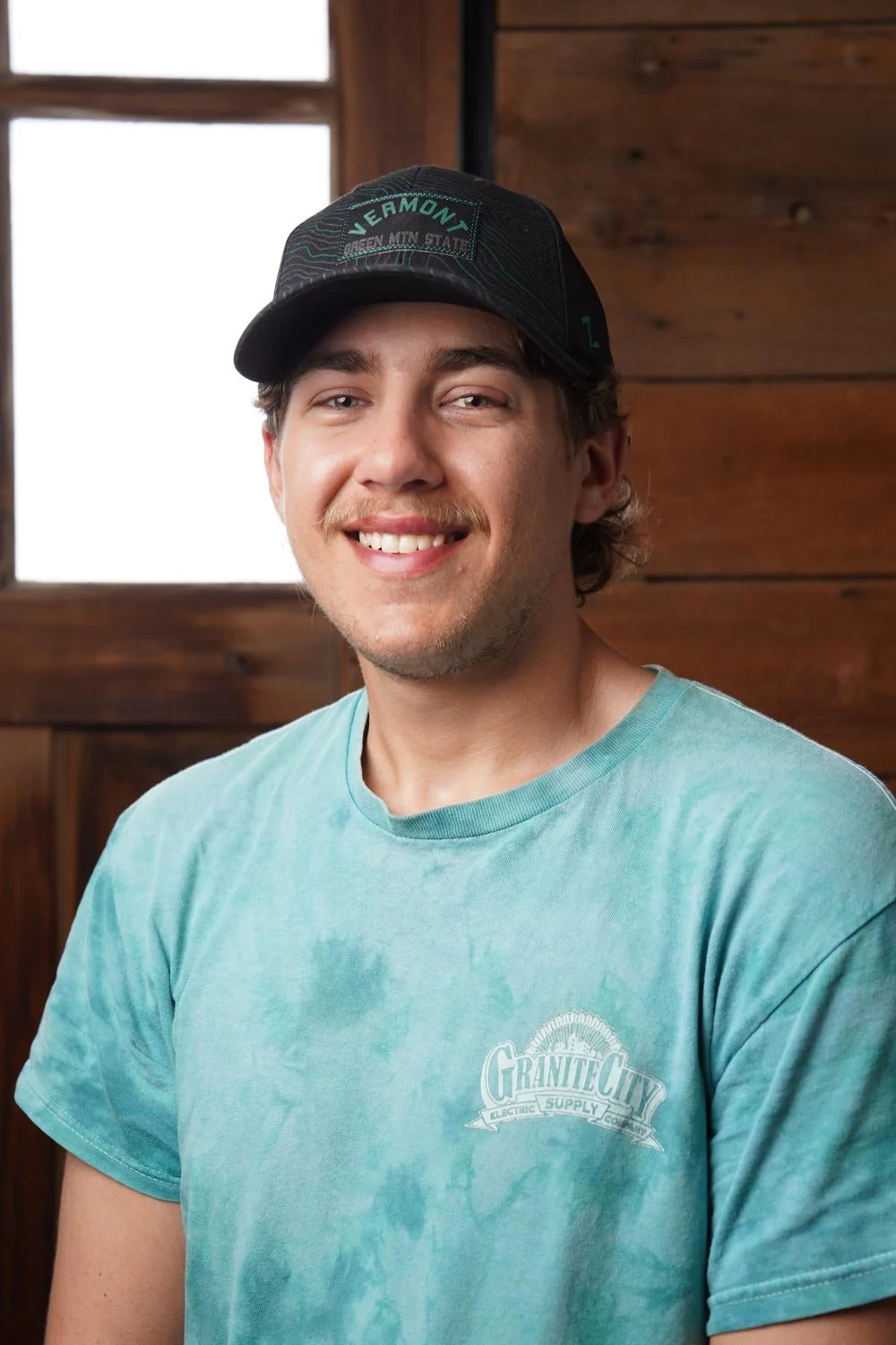 A smiling young man with light brown hair and facial hair, wearing a turquoise T-shirt with a "Granite City" logo, standing indoors with a background of wood paneling and a window.