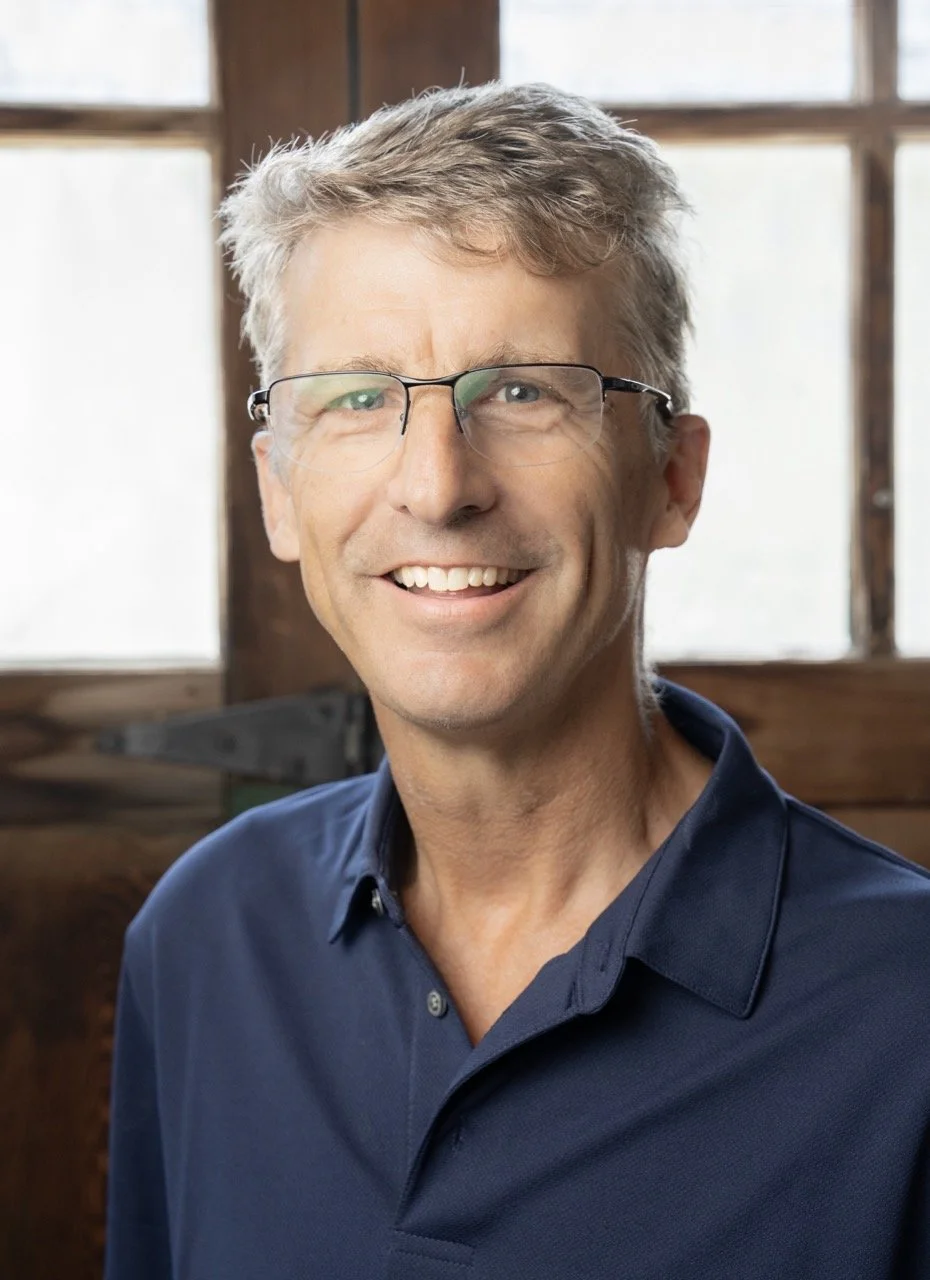 Tim Carruthers with short gray hair smiling outdoors, wearing a gray outdoor jacket over a checkered shirt, with trees and grass in the background.