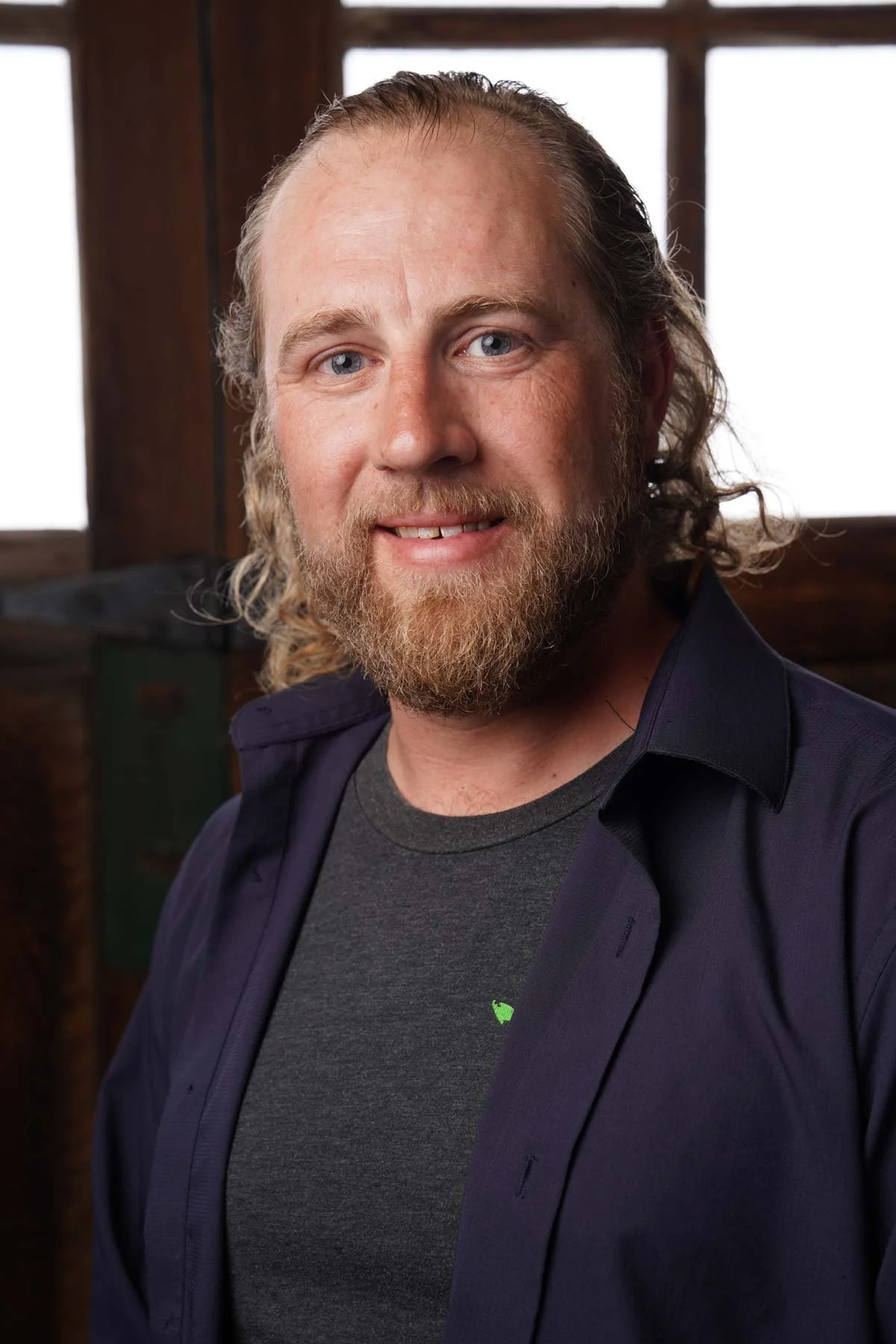 A man with curly hair and a beard wearing a striped shirt and glasses resting on his head, sitting on a wooden bench against a wooden shingle wall.