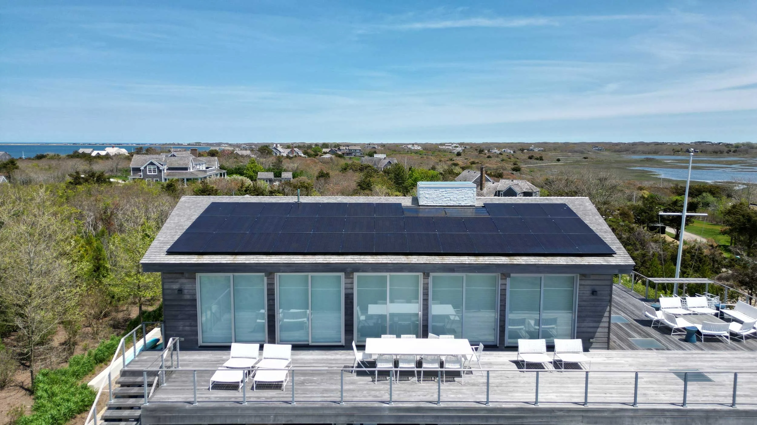 A house with solar panels on its roof, situated on a coastal landscape with many other houses, trees, and water in the background, under a partly cloudy sky.