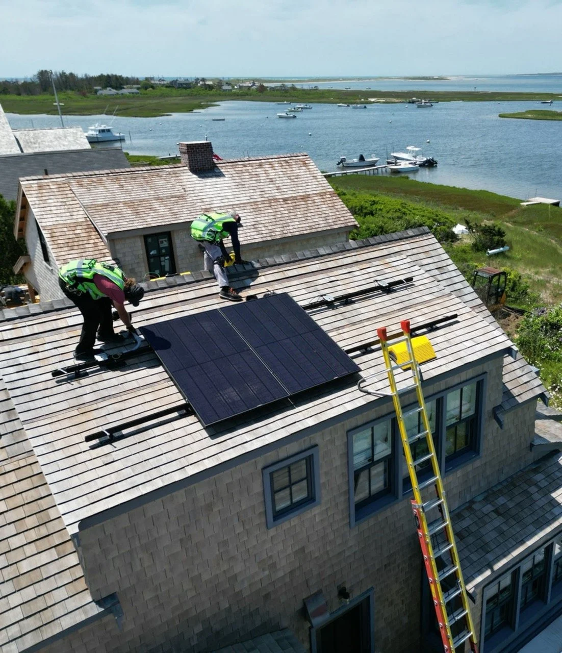 Three workers installing solar panels on the roof of a house overlooking a body of water with boats.