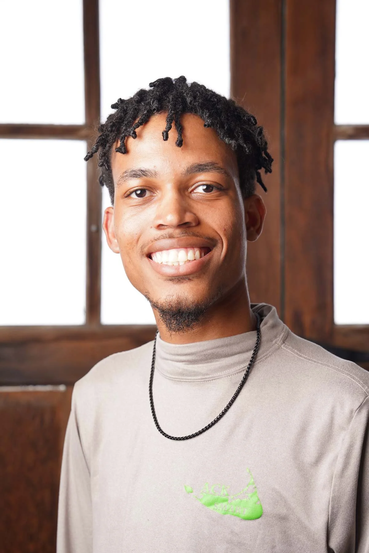 Young man smiling, wearing a light-colored t-shirt with a green logo, black beaded necklace, standing in front of a wooden-paneled window with white background.