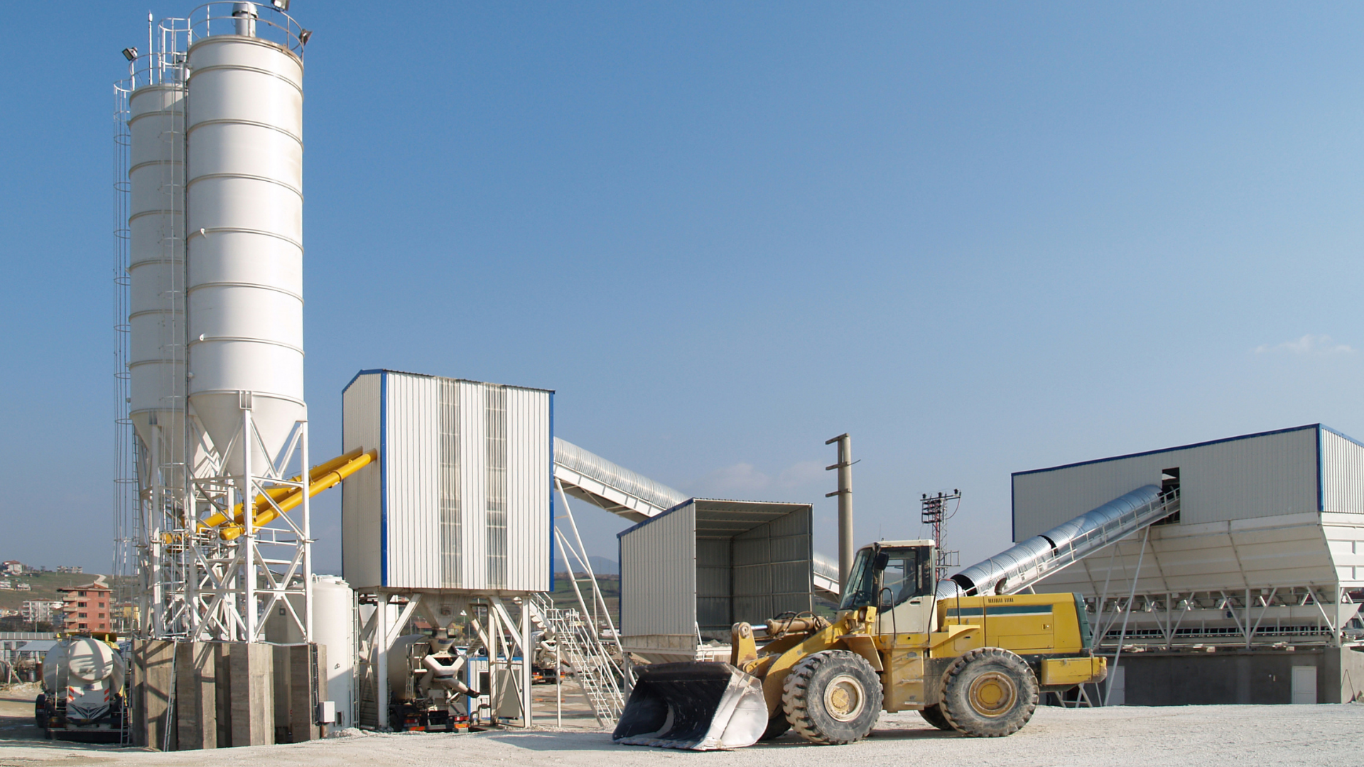 Fábrica de cemento con silos grandes, estructuras metálicas, maquinaria y un bulldozer en primer plano bajo un cielo despejado.