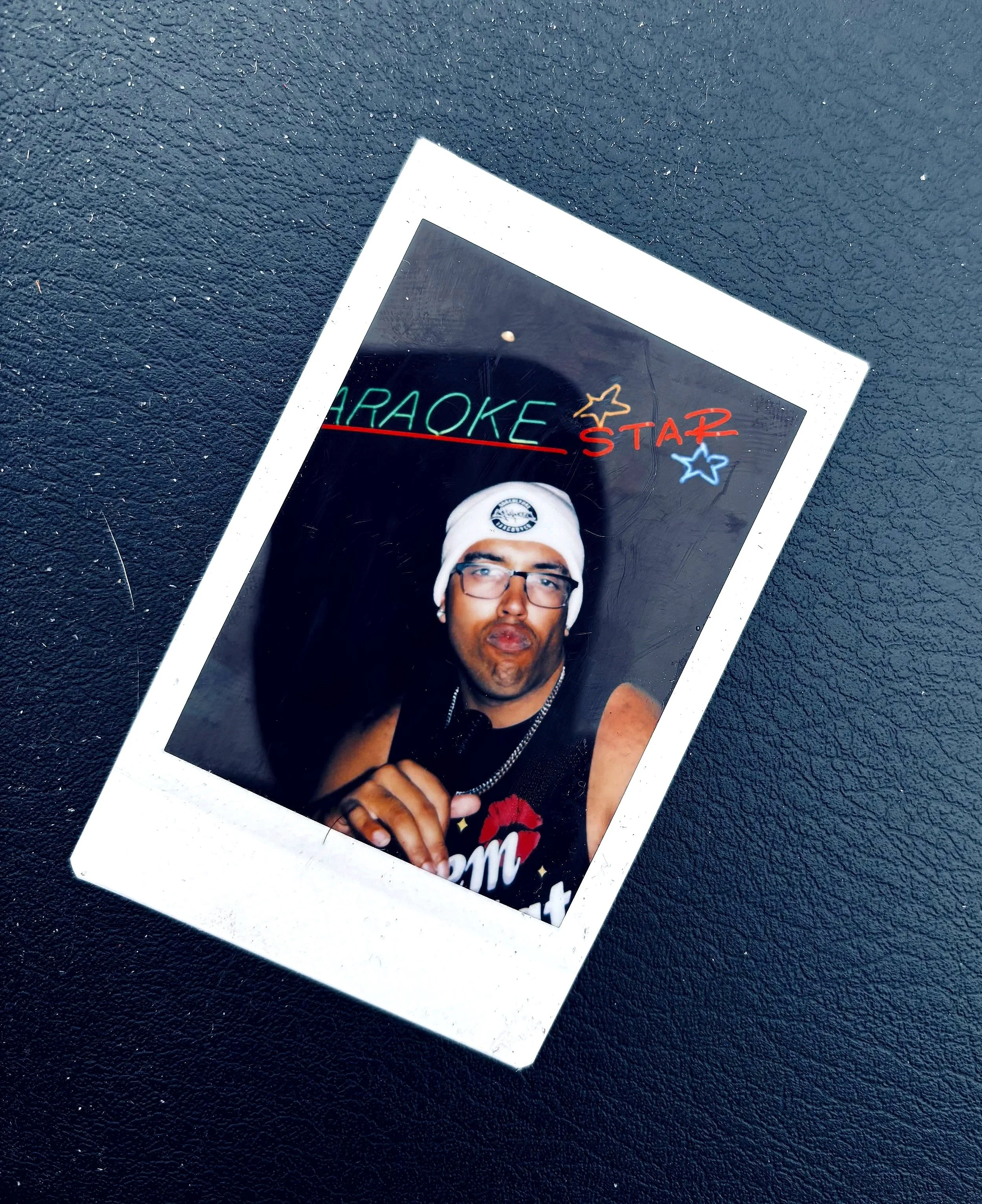 Photograph of a person with a beard, glasses, and a white knit hat taking a selfie. The photo appears on a small printed card lying on a dark textured surface. The card has colorful drawings and text, including the words 'KARAOKE STAR' with stars around it.