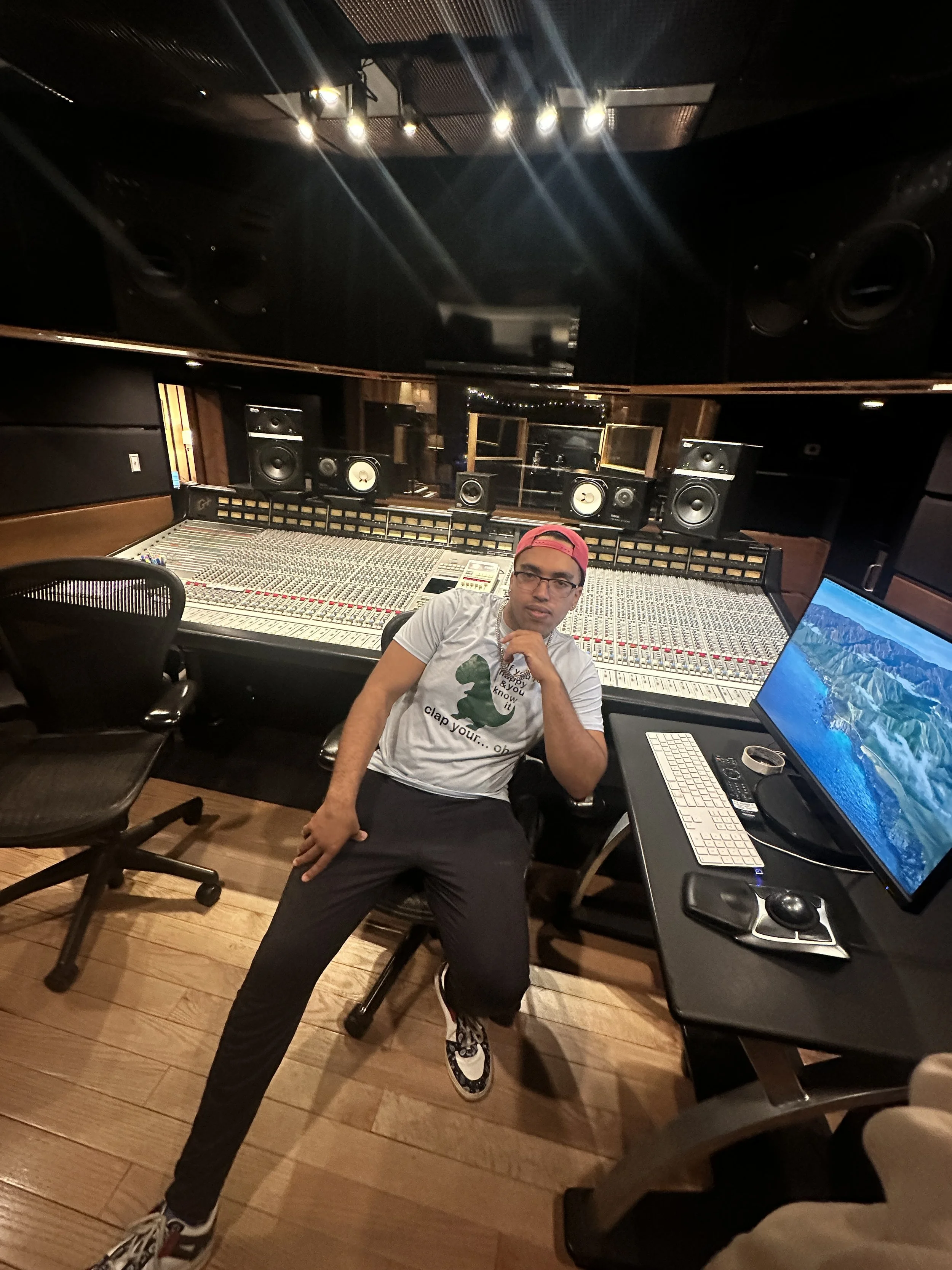 A man wearing glasses, a backwards red baseball cap, a t-shirt with a dinosaur, and black pants, sitting in a music recording studio with a large mixing console and various studio speakers, looking at the camera.
