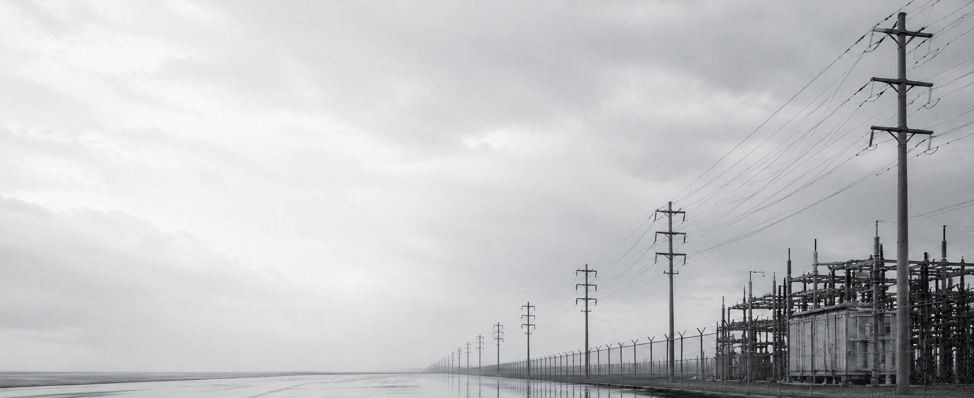 Overcast sky over a long row of electrical power lines and electric substation next to a chain-link fence along a coastal area.