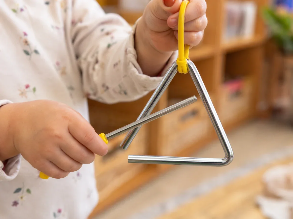 Child holding a metal triangle musical instrument with a yellow striker in a room with wooden shelves.