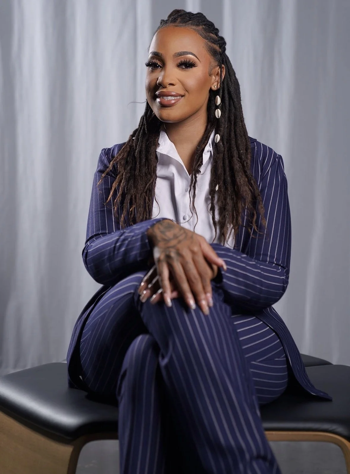 A woman with dreadlocks and tattoos, dressed in a navy blue pinstripe suit and white shirt, sitting on a black bench with a gray curtain background.
